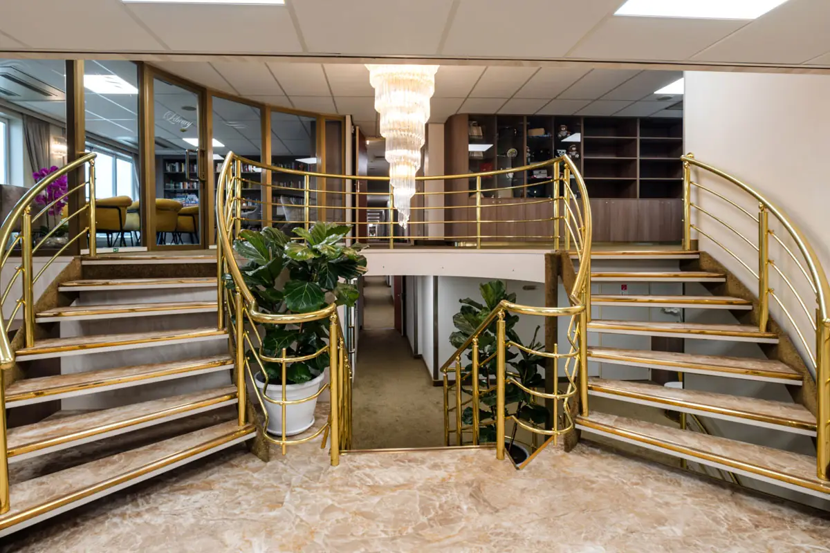 Doppelte Treppe mit goldfarbenem Geländer in einer Lobby, mittig große Pflanze und Kronleuchter über dem Treppenabsatz