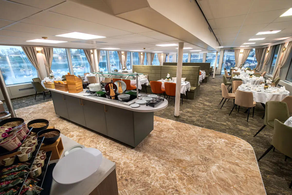 Speisesaal mit gedeckten Tischen, Stühlen und Buffetbereich, große Fenster mit Vorhängen bieten Blick nach draußen