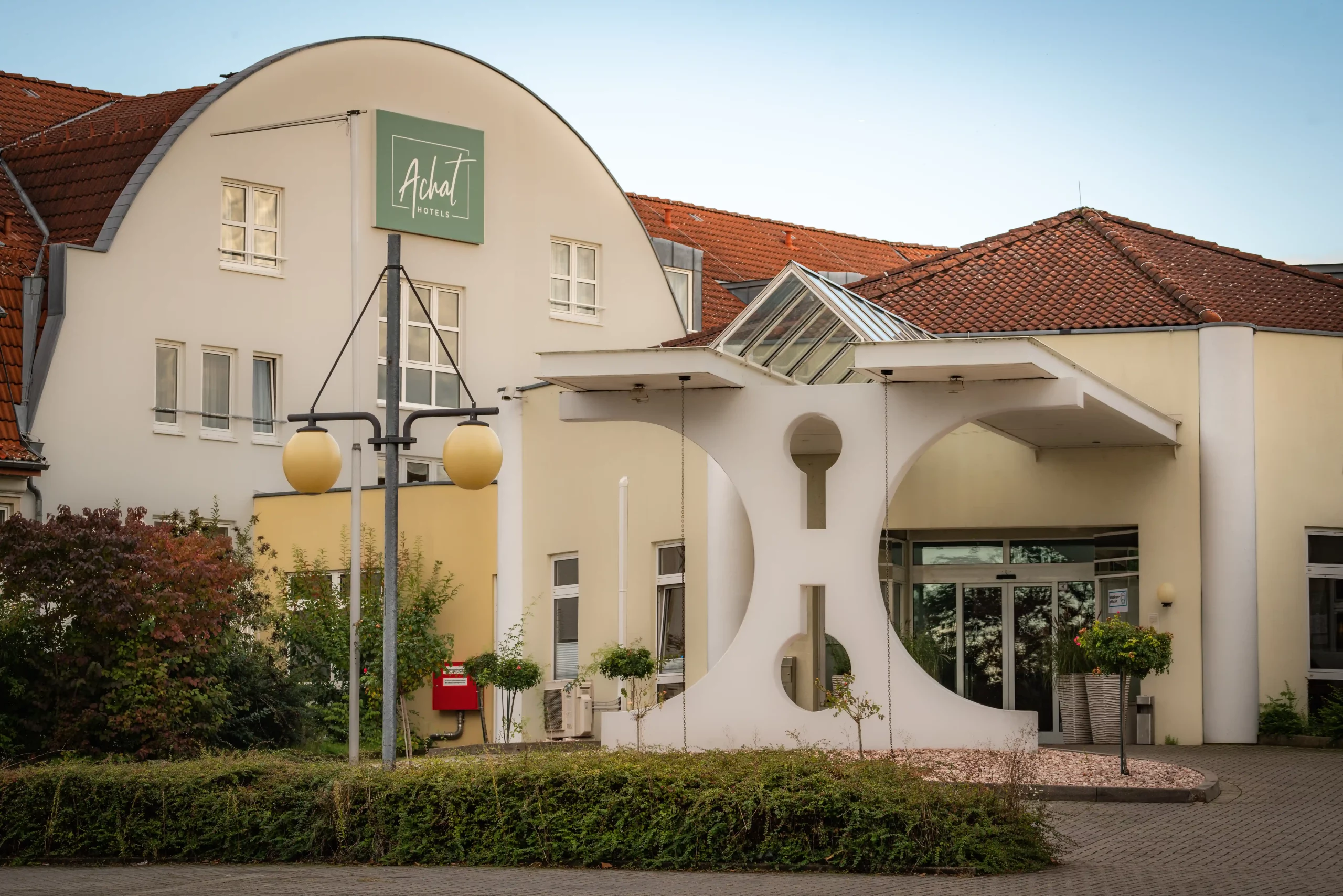 Eingangsbereich des ACHAT Hotels Reilingen Walldorf mit rundem Vordach, zwei Laternen und grünem Schild mit weißer Aufschrift