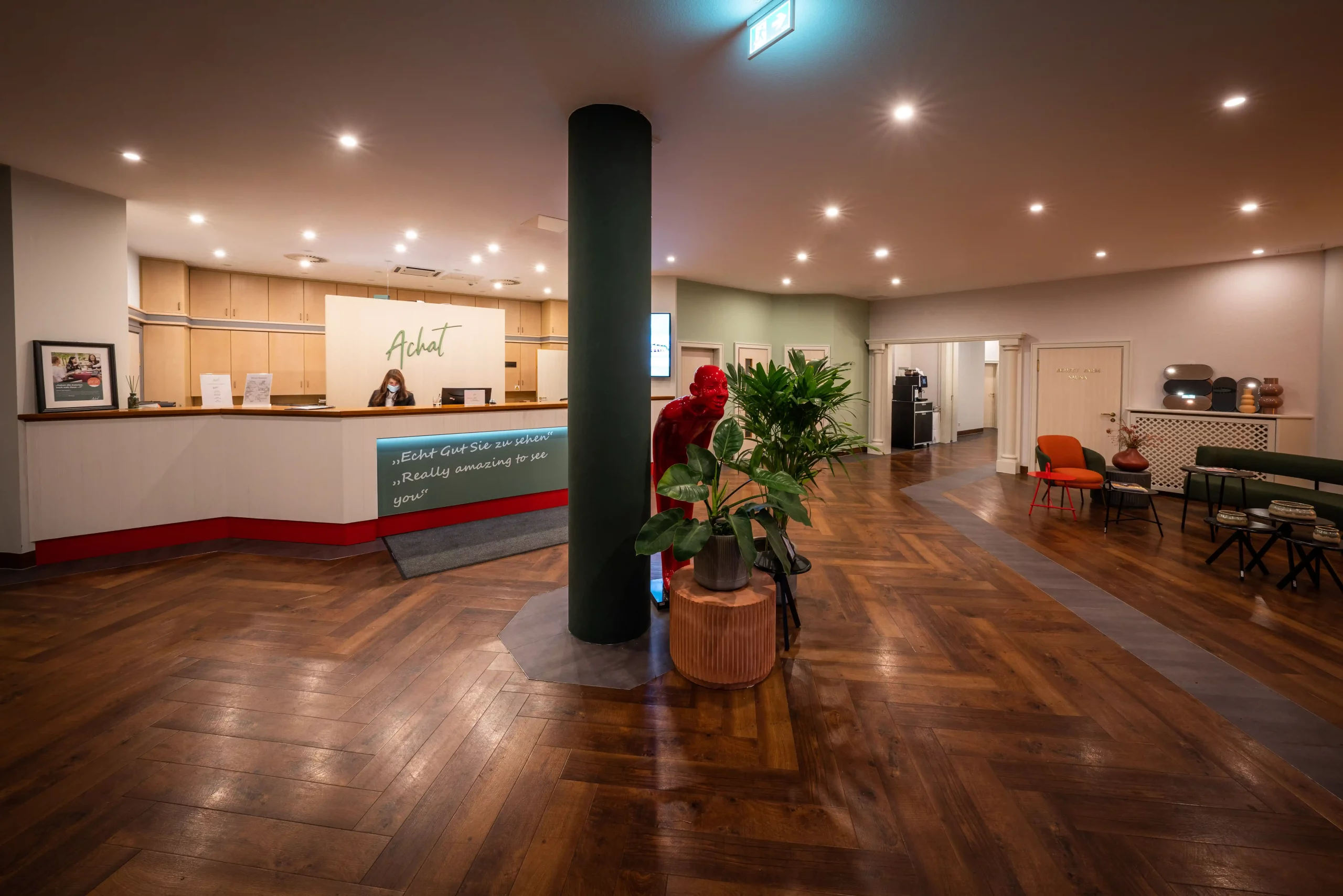 Empfangsbereich des ACHAT Hotels Reilingen Walldorf mit Rezeption, zwei Personen hinter dem Tresen, großem schwarzen Säulenpfeiler in der Mitte, Pflanzen und Sitzgelegenheiten mit Stühlen und Tischen auf dunklem Parkettboden.