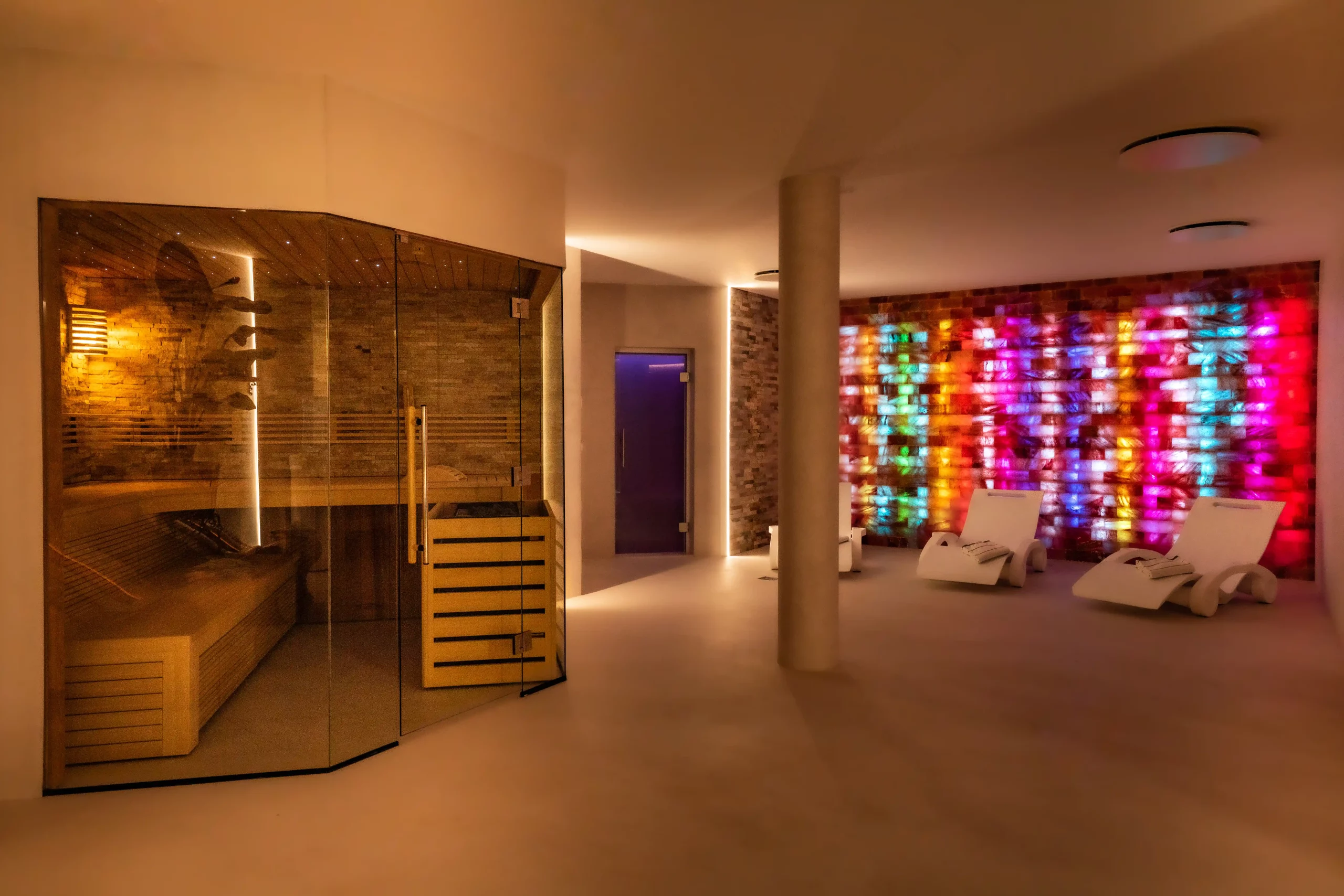 Moderner Wellnessbereich mit Sauna aus Glas und Holz links sowie zwei weißen Liegen vor einer farbig beleuchteten Wand aus Glasbausteinen rechts