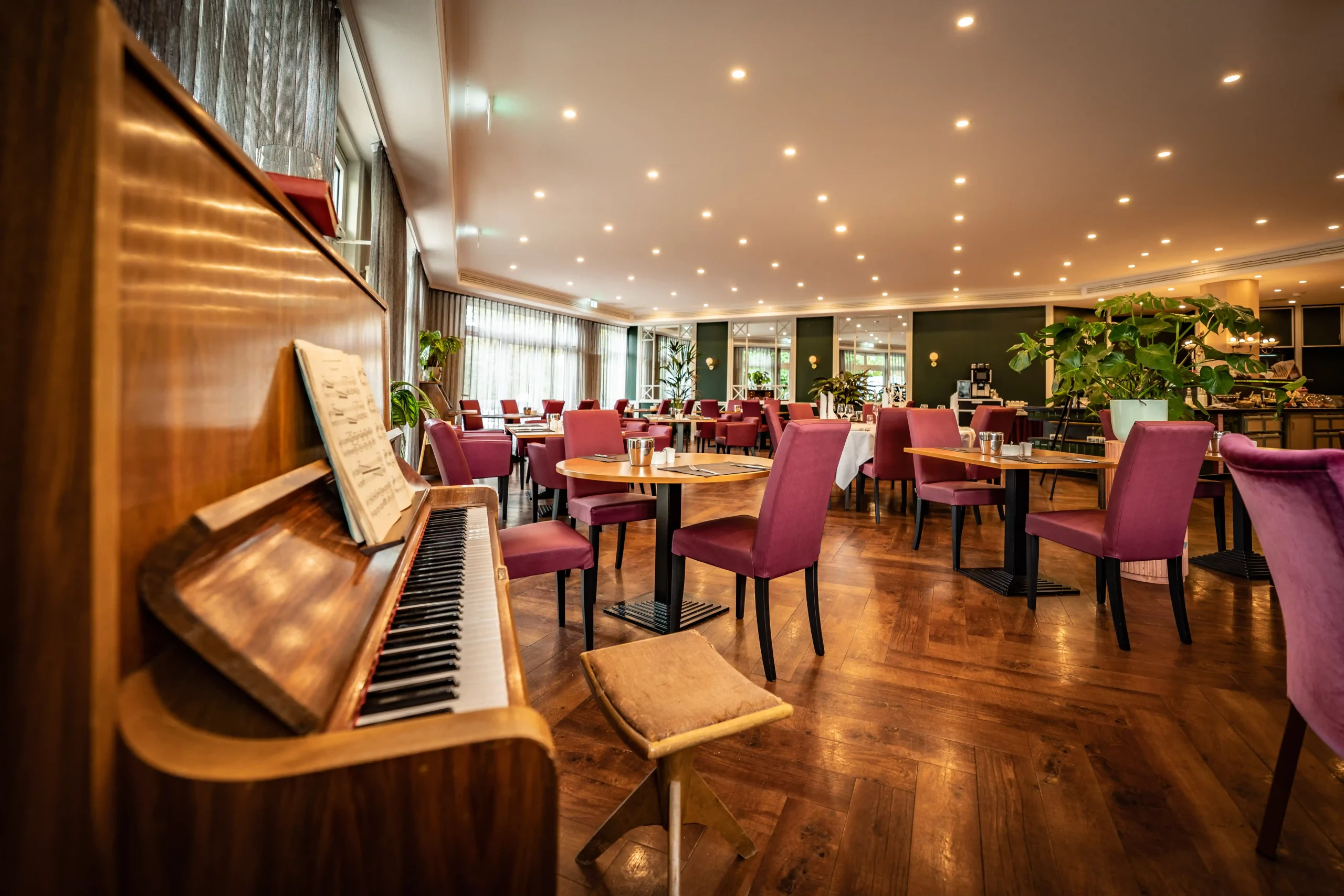 Restaurant mit Holzboden, mehreren runden Tischen und Stühlen mit roten Polstern, links ein aufgeschlagenes Klavier mit Hocker, große Fenster mit Vorhängen und viele Deckenlampen.