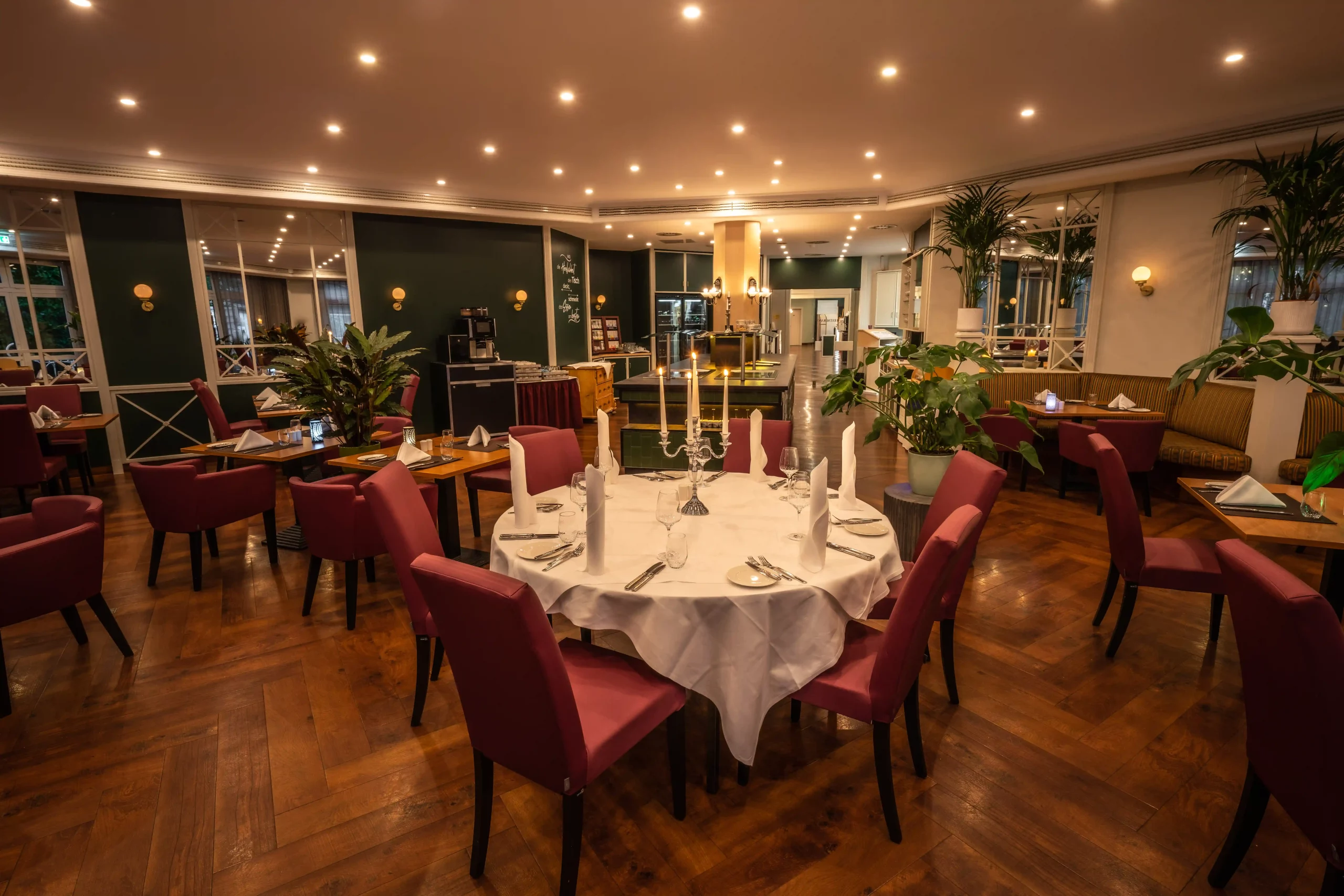 Rund gedeckter Tisch mit weißer Tischdecke, Kerzenleuchter und roten Stühlen in einem warm beleuchteten Restaurant mit Holzboden und weiteren Tischen im Hintergrund.