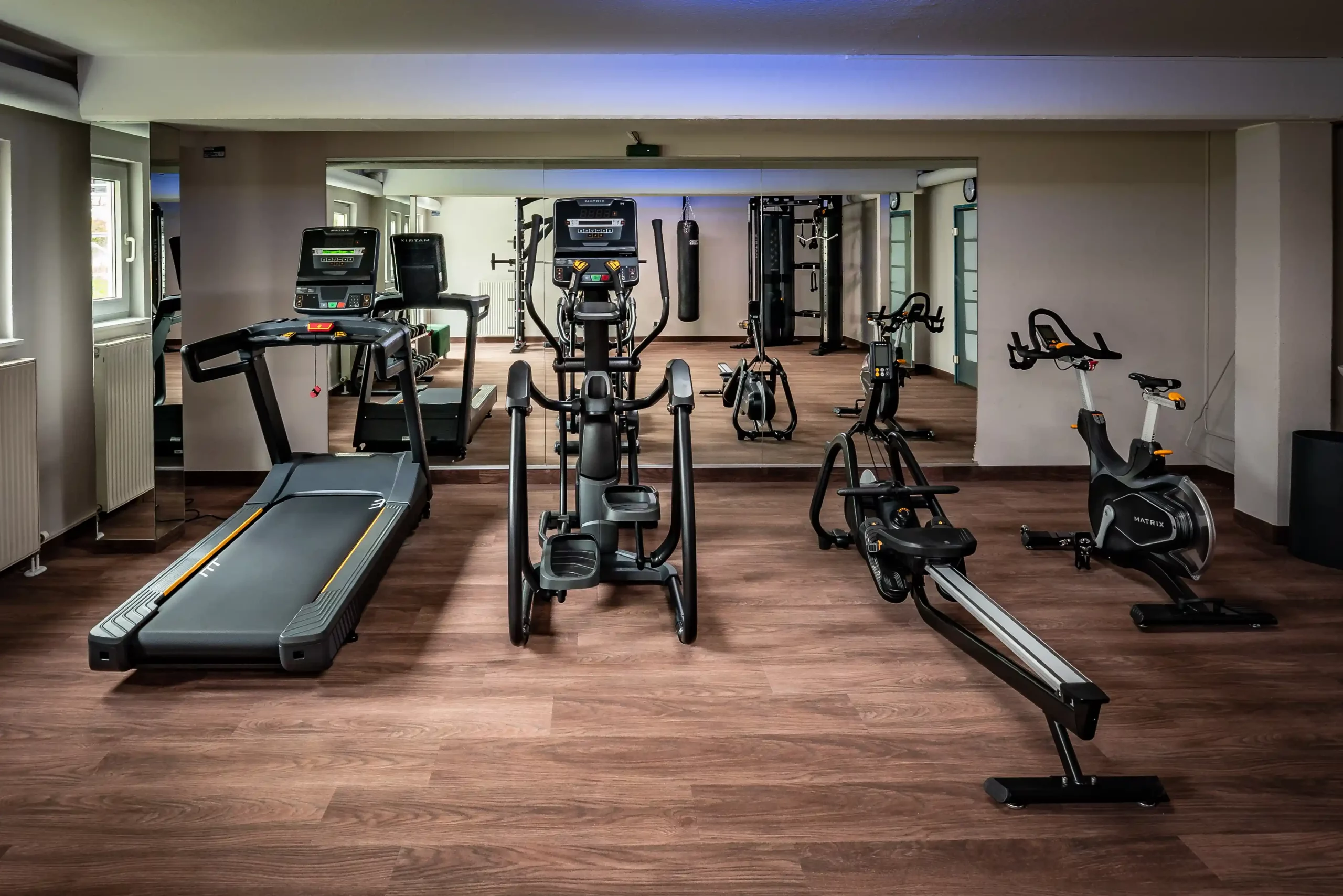 Fitnessraum mit Laufband, zwei Crosstrainern, Rudergerät, Spinningbike und Kraftstation vor großen Spiegeln und Holzfußboden im ACHAT Hotel Reilingen Walldorf.