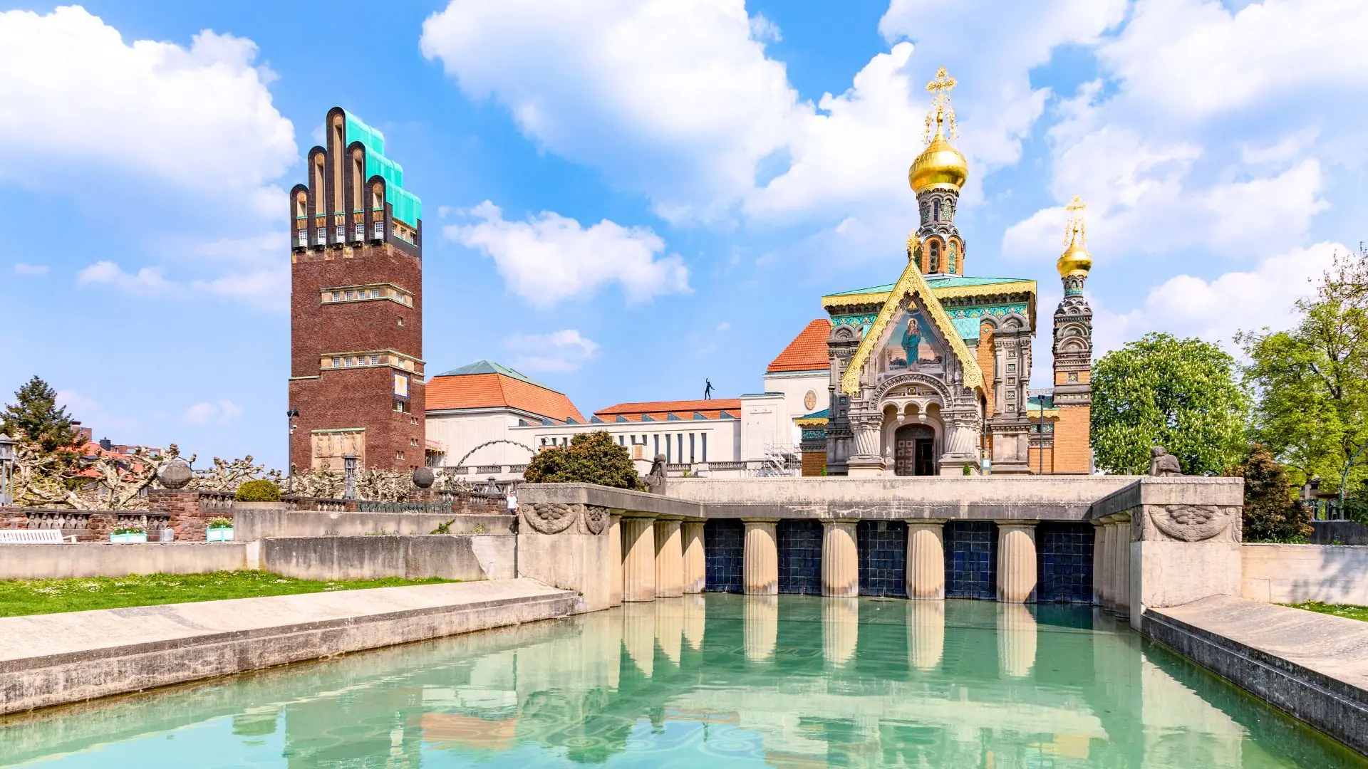 Blick auf den Hochzeitsturm und die Russische Kapelle in Darmstadt mit einem Wasserbecken im Vordergrund und Bäumen im Hintergrund