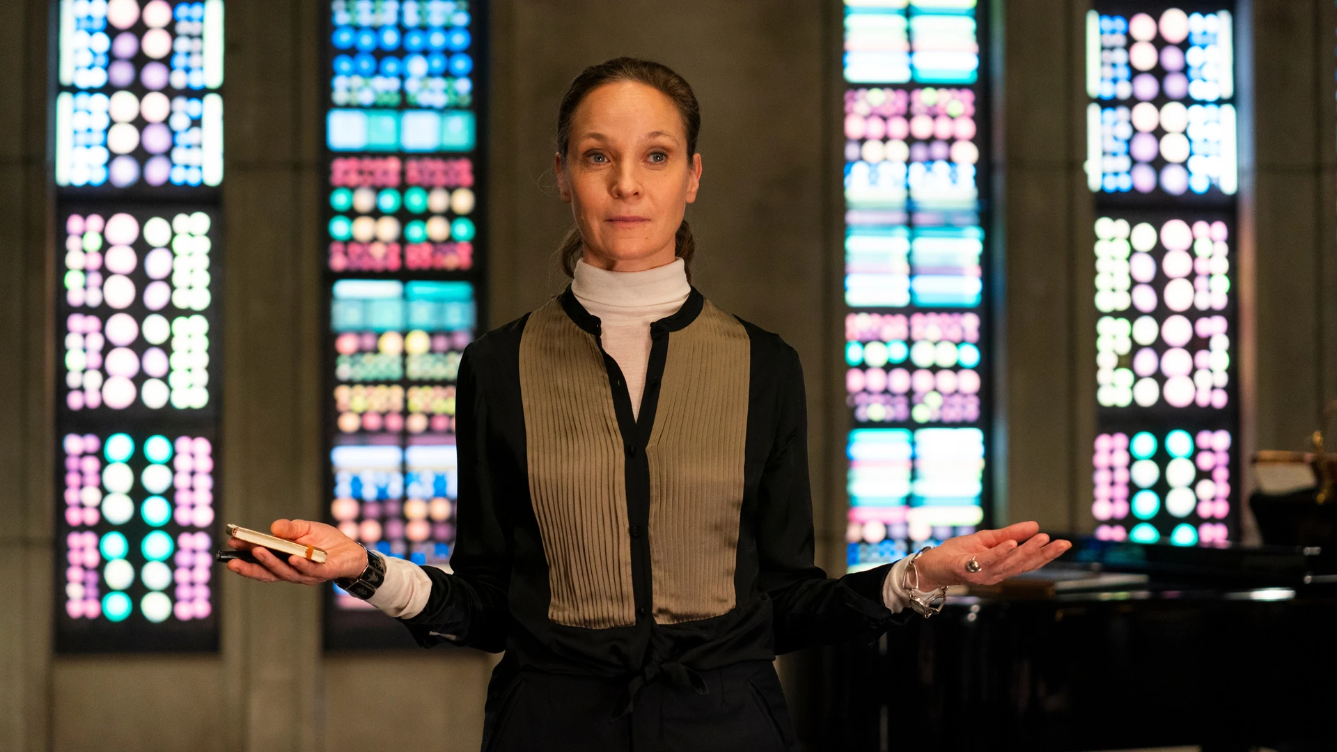 Regina Feldmann (Jeanette Hain) steht mit geöffneten Armen vor bunten Fenstern. Im Hintergrund steht ein Flügel.