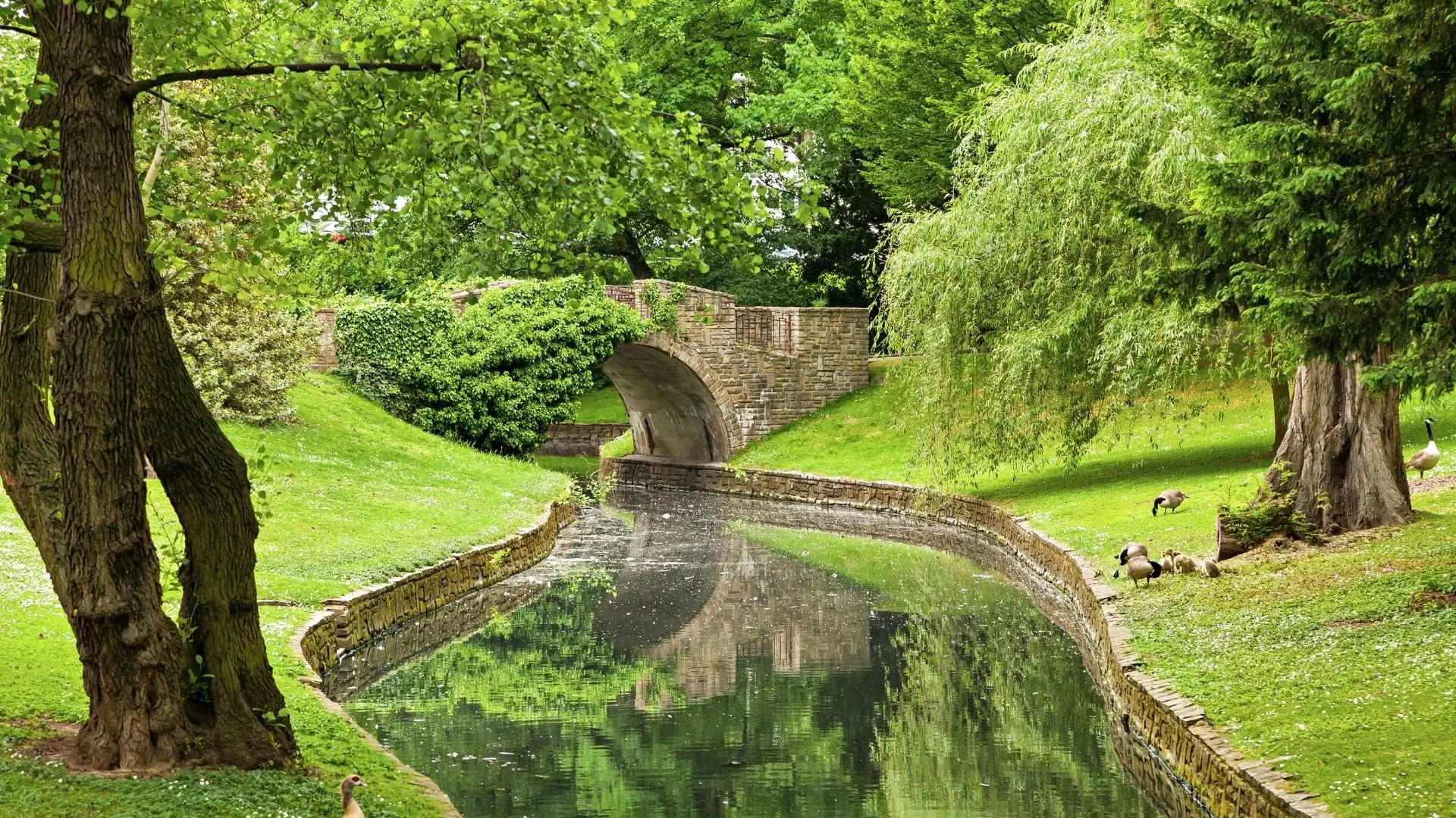 Blick auf einen gewundenen Wasserlauf im Park der Boverie mit einer steinernen Brücke und umgebender grüner Vegetation.