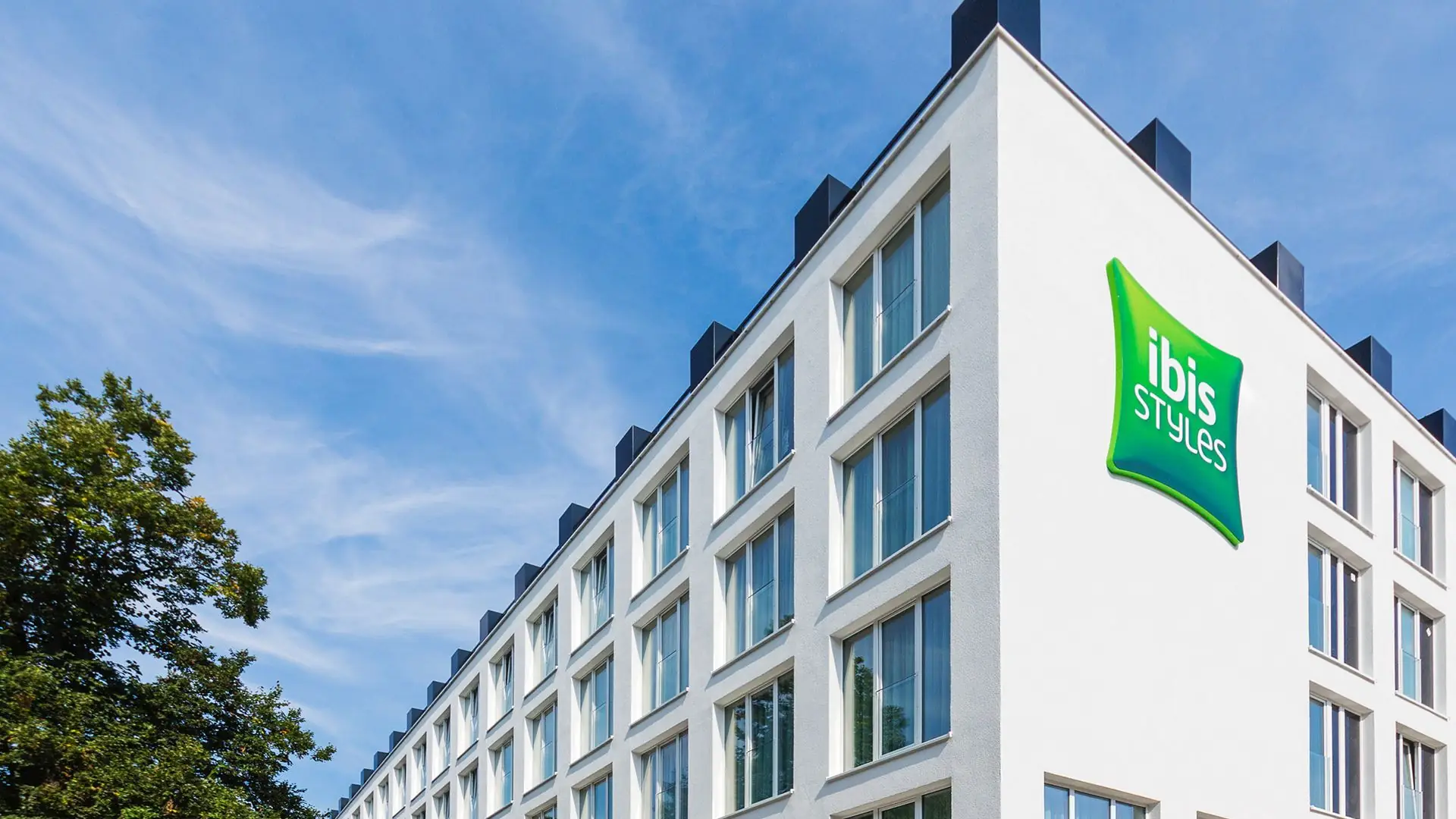 Mehrstöckiges weißes Hotelgebäude mit vielen Fenstern und grünem ibis Styles Logo an der Ecke