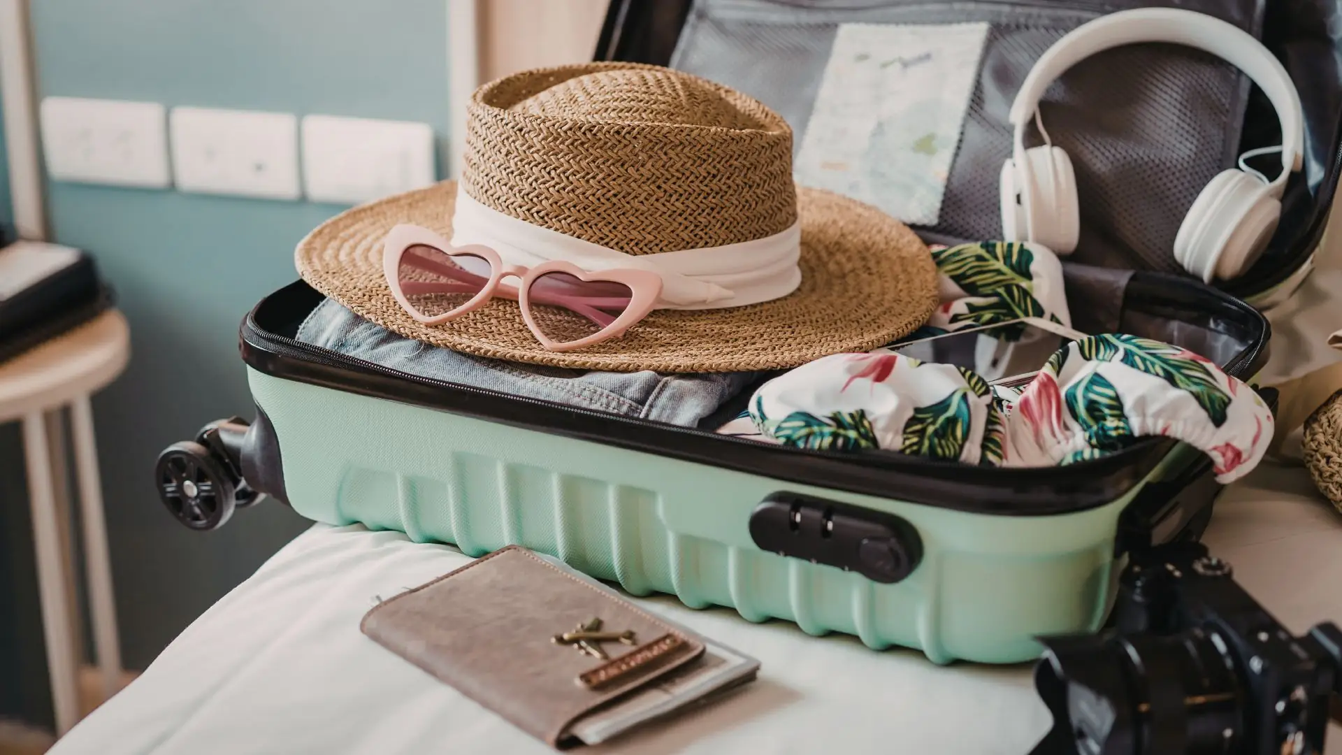 Offener, mintgrüner Koffer mit Strohhut, herzförmiger Sonnenbrille, Bikini und weißen Kopfhörern auf einem Bett