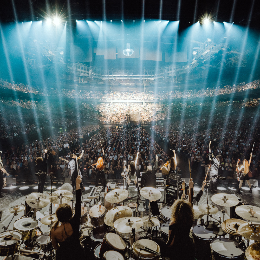 Hans Zimmer und seine Band während einer Live-Performance vor einem beeindruckenden, vollbesetzten Publikum. Die Bühne ist von strahlenden Lichtkegeln durchflutet, die eine mitreißende Atmosphäre schaffen und die Energie des Konzerts unterstreichen.