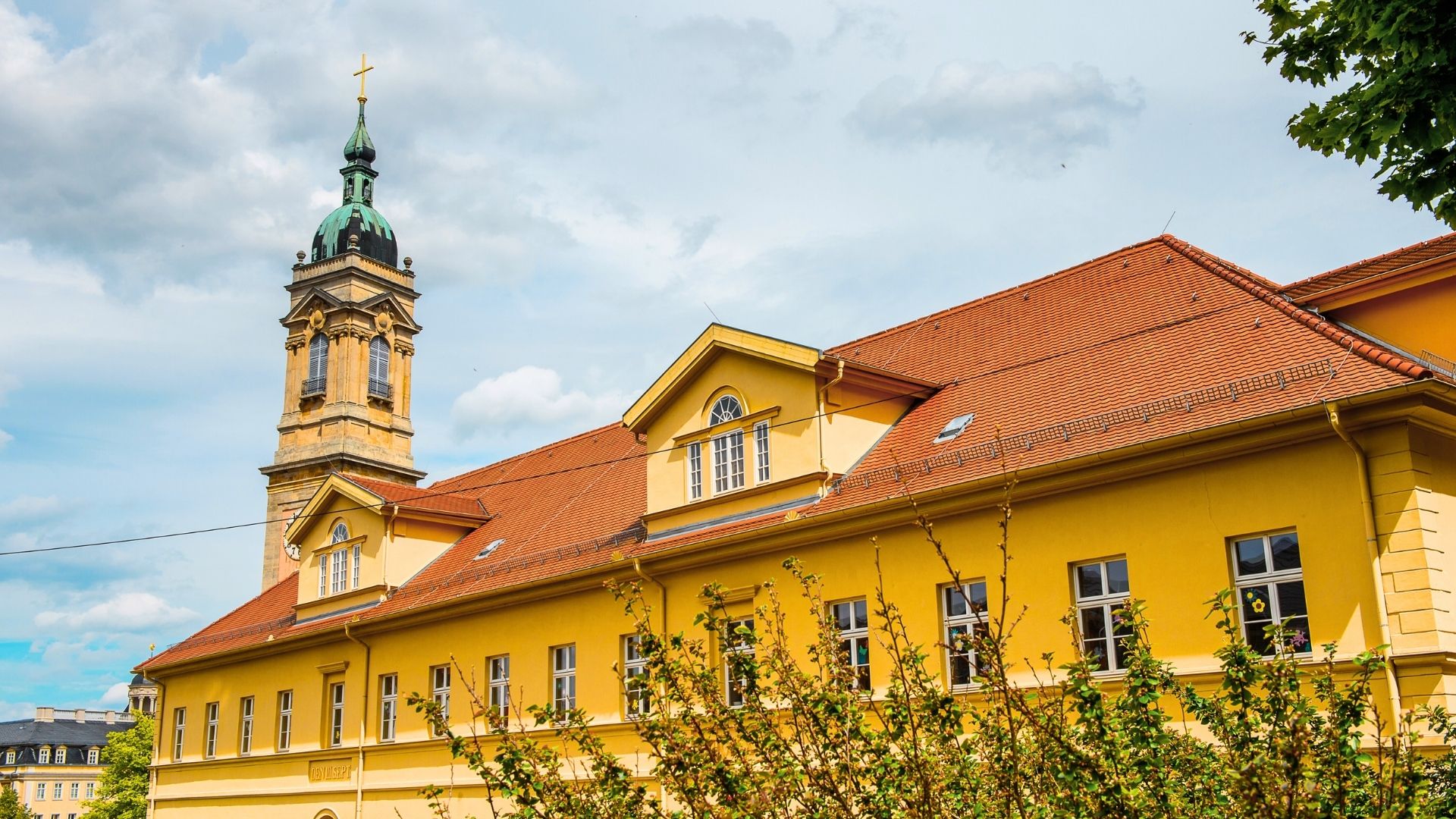 Ansicht der Georgenkirche in Eisenach mit einem hohen Turm und einer goldenen Kreuzspitze. Das Gebäude hat eine gelbe Fassade und ein rotes Dach.