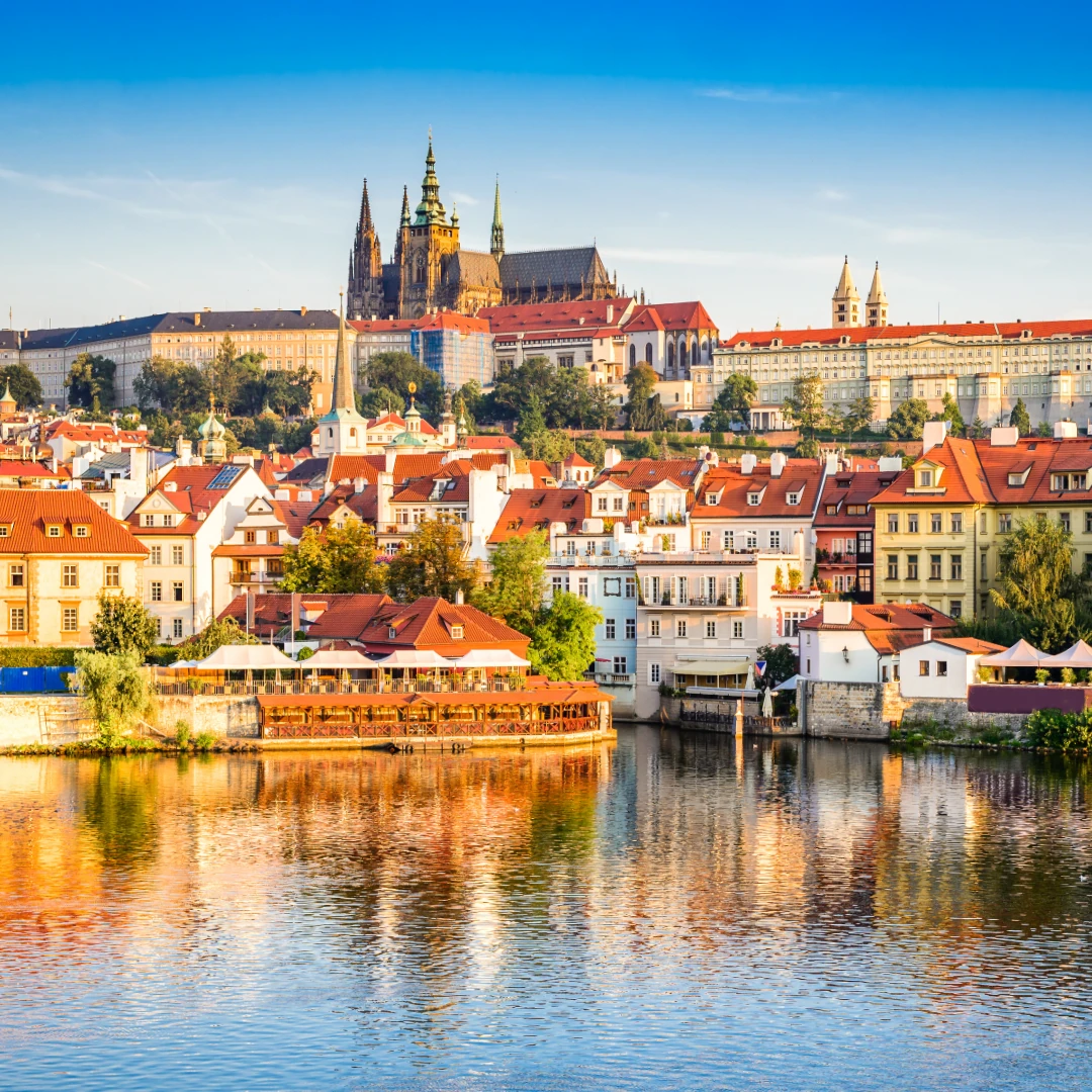 Dieses Foto zeigt die malerische Skyline von Prag mit der Prager Burg im Hintergrund. Die roten Dächer der historischen Altstadt spiegeln sich im ruhigen Wasser der Moldau und verleihen der Stadt einen charmanten und romantischen Charakter.