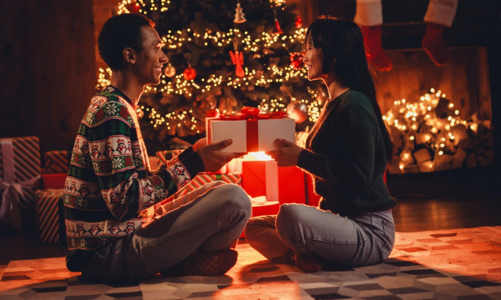 Das Bild zeigt ein Paar, das sich an Weihnachten gegenseitig ein Geschenk überreicht. Beide lächeln sich an, während sie sich liebevoll anschauen. Der Mann trägt einen blauen Pullover, die Frau ein rotes Kleid. Im Hintergrund ist ein festlich geschmückter Weihnachtsbaum mit Geschenken zu sehen. Die Atmosphäre ist warm und einladend, was eine gemütliche und fröhliche Weihnachtsstimmung vermittelt.
