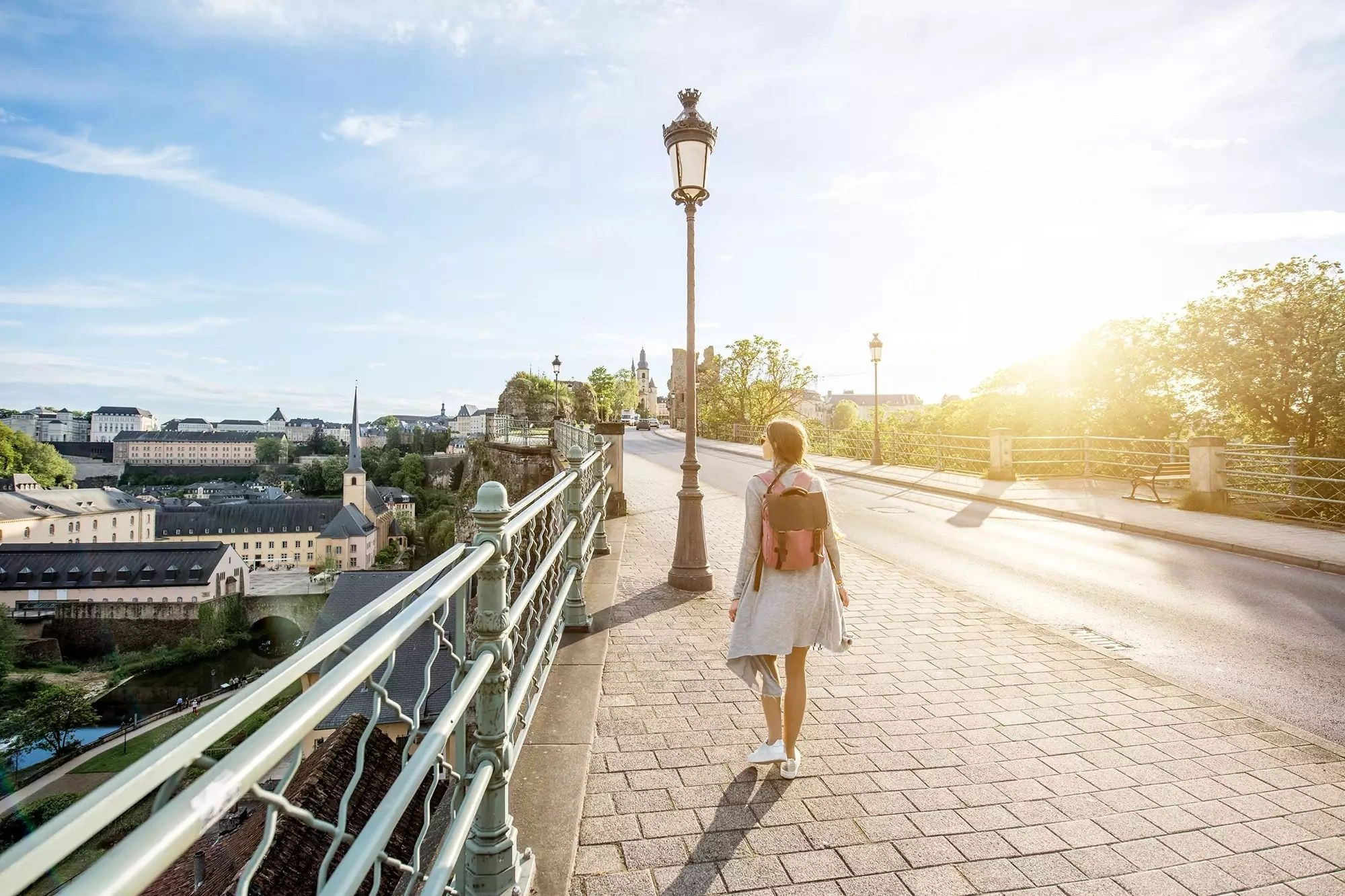 Eine Touristin beim Spaziergang über eine Brücke während des Sonnenaufgangs in Luxemburg, mit Blick auf die malerische Stadt Luxemburg. Die sanften Lichtstrahlen des Morgens verleihen der Szene eine zauberhafte Atmosphäre.