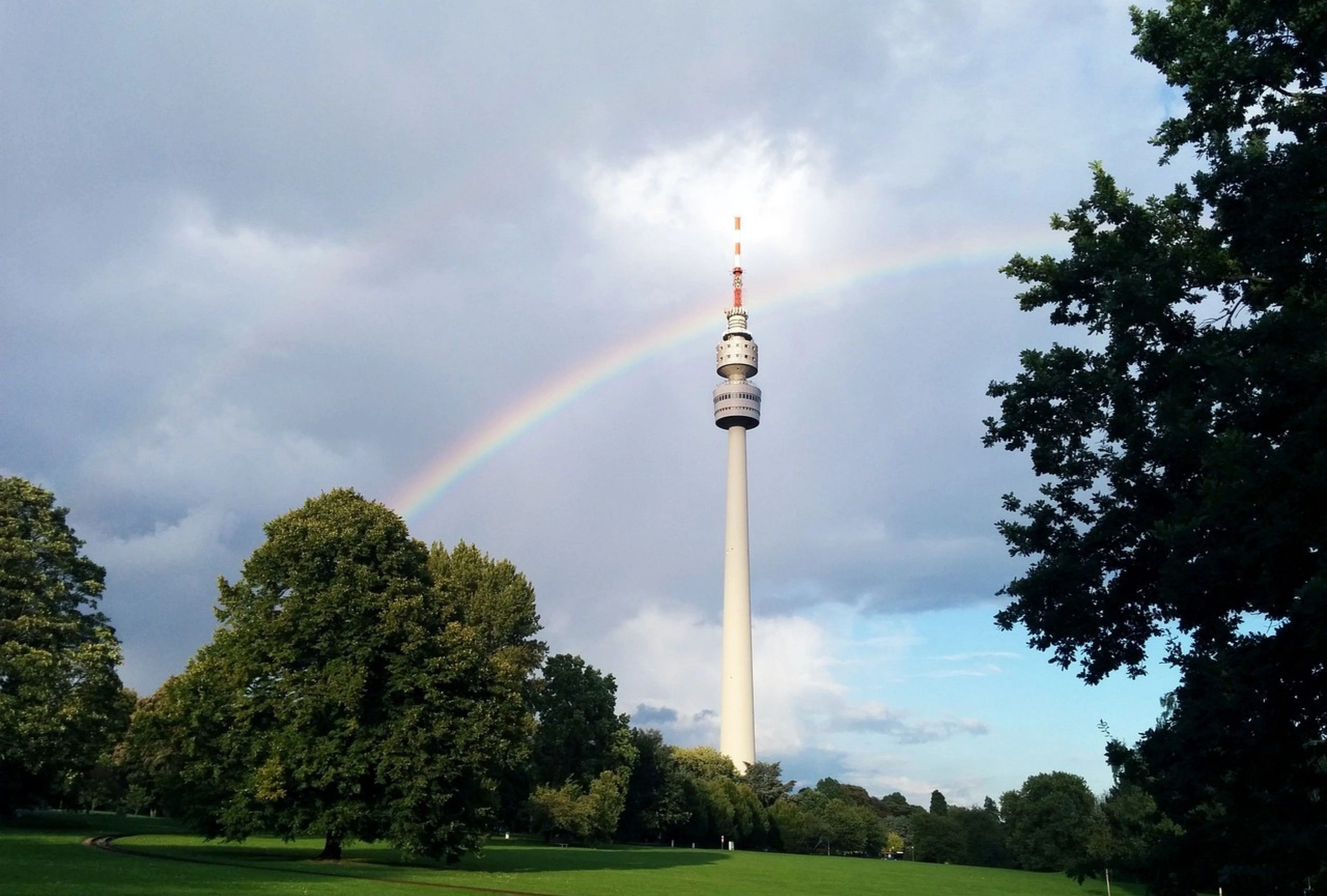 Fernsehturm in einem Park mit grünen Bäumen und einem Regenbogen am bewölkten Himmel