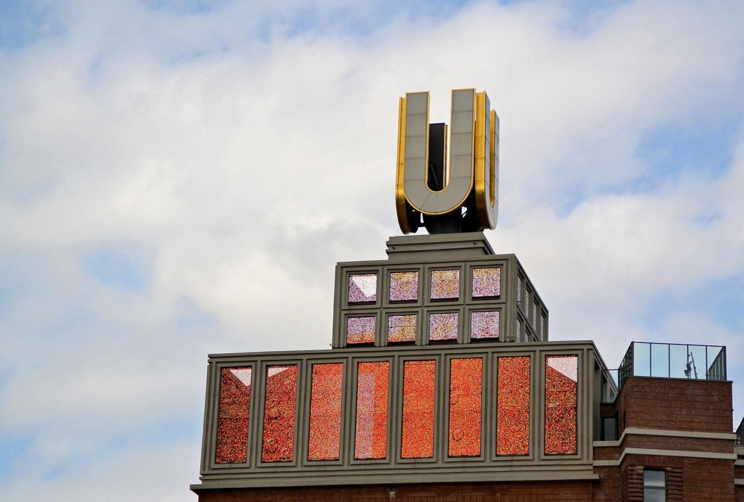 Dach eines Gebäudes mit großem goldfarbenem Buchstaben 'U' auf einem grauen Sockel, darunter Fenster mit roten und violetten Glaselementen, bewölkter Himmel im Hintergrund