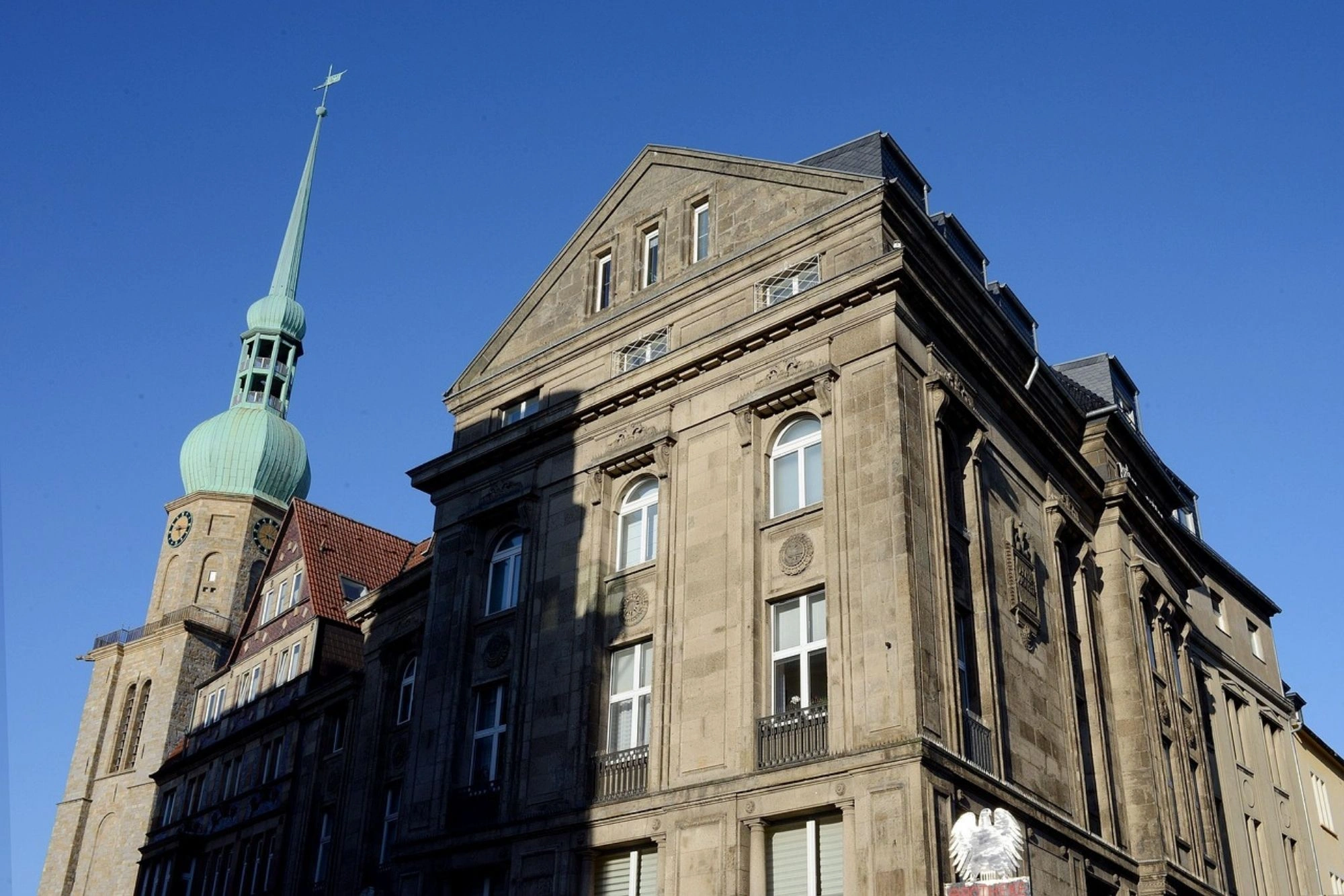 Historisches Gebäude mit Sandsteinfassade und mehreren Fenstern neben einem Kirchturm mit grünem, spitzem Dach und Kreuz