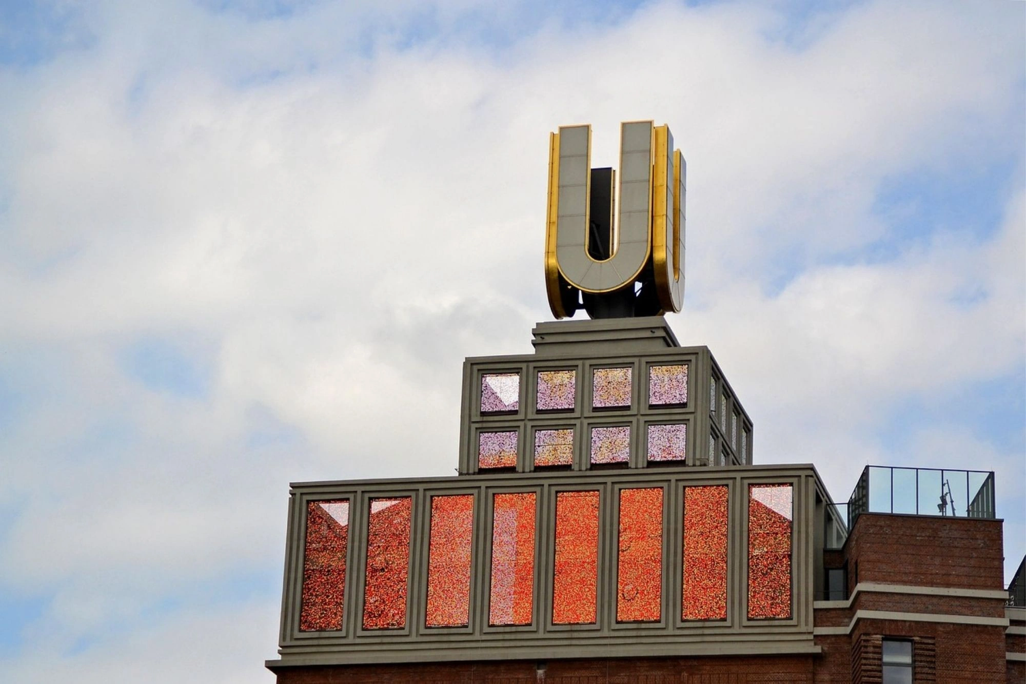 Dach eines Gebäudes mit großem goldfarbenem Buchstaben 'U' auf einem grauen Sockel, darunter Fenster mit roten und violetten Glaselementen, bewölkter Himmel im Hintergrund