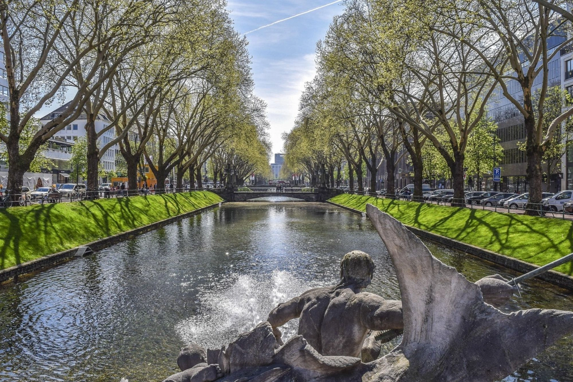 Blick auf einen von Bäumen gesäumten Kanal mit einer Brücke im Hintergrund und einer Wasserskulptur im Vordergrund