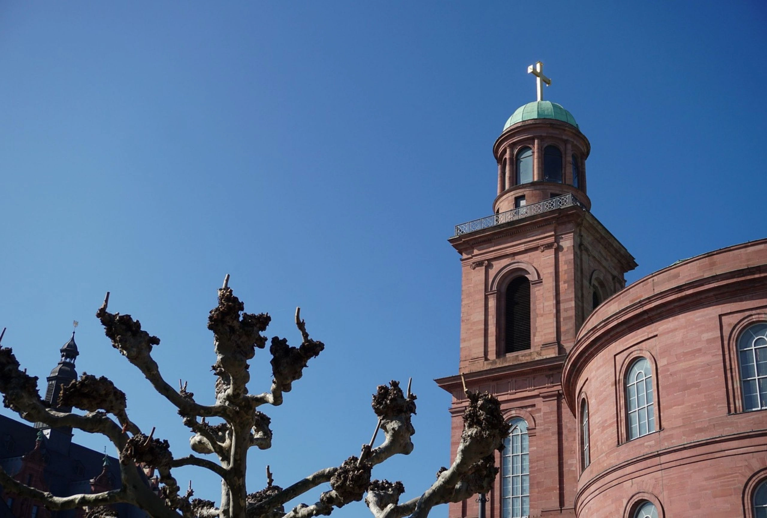Kirchturm mit grünem Kuppeldach und goldenem Kreuz vor blauem Himmel, im Vordergrund knorrige Baumäste