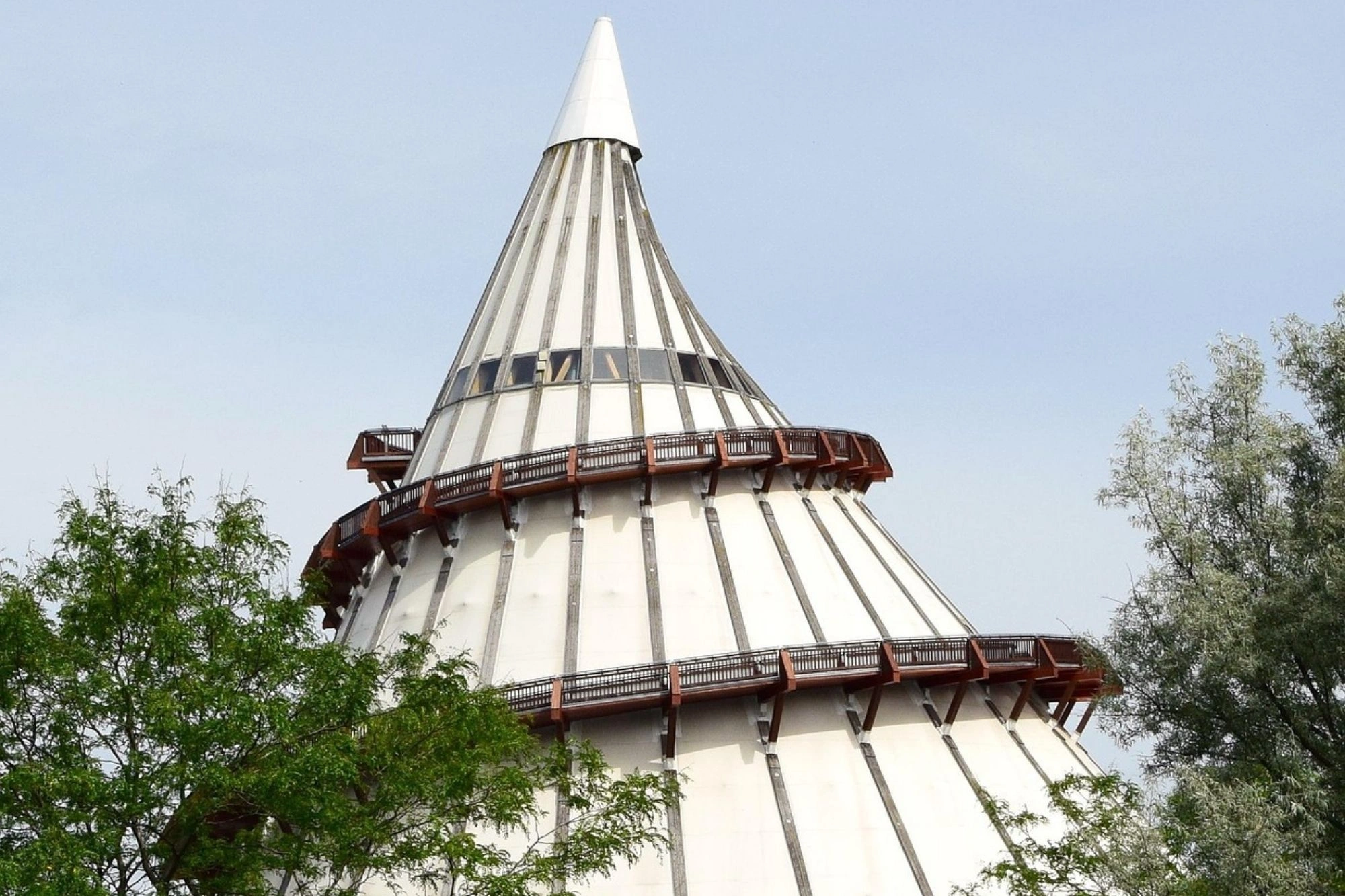 Spitz zulaufender Turm mit zwei umlaufenden Balkonen, umgeben von Bäumen