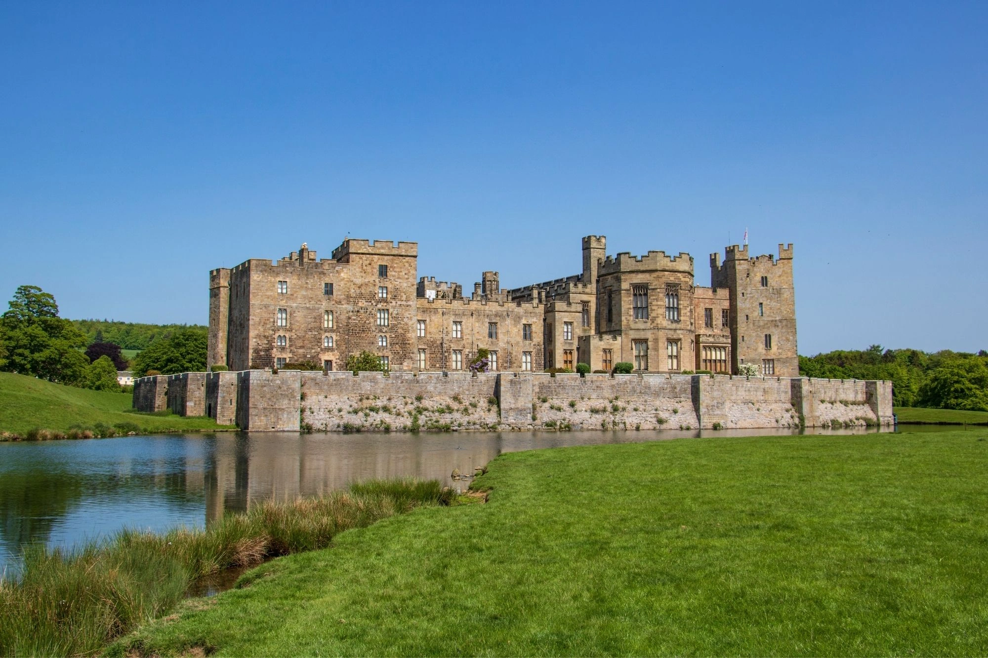 Historisches Schloss mit Wehrmauern und Türmen an einem Seeufer, umgeben von grüner Wiese und blauem Himmel
