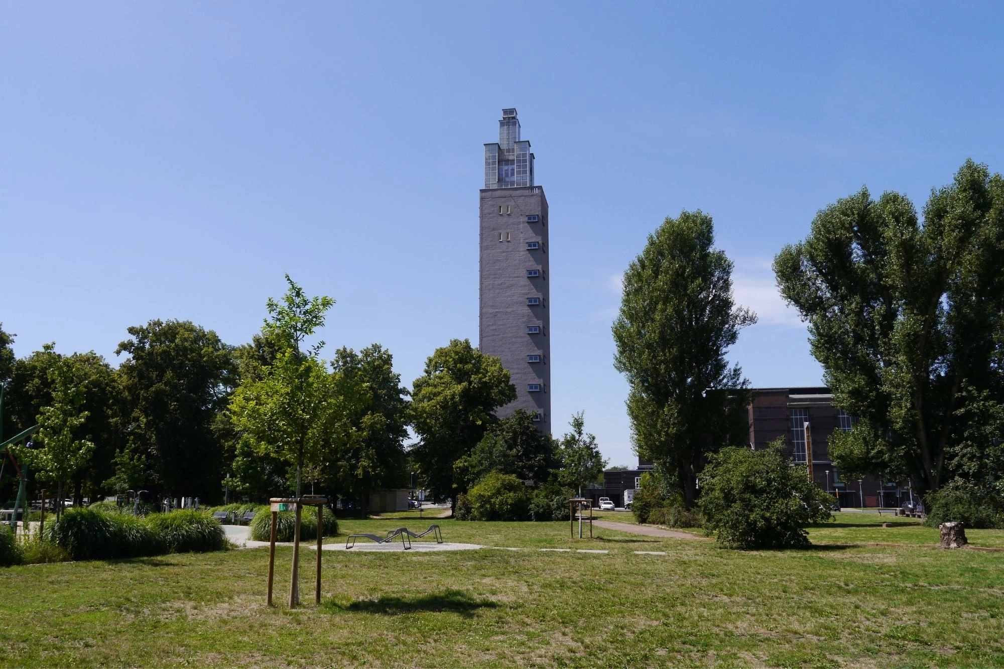 Hoher, schmaler Turm aus Beton mit mehreren kleinen Fenstern, umgeben von Bäumen und einer Wiese im Vordergrund