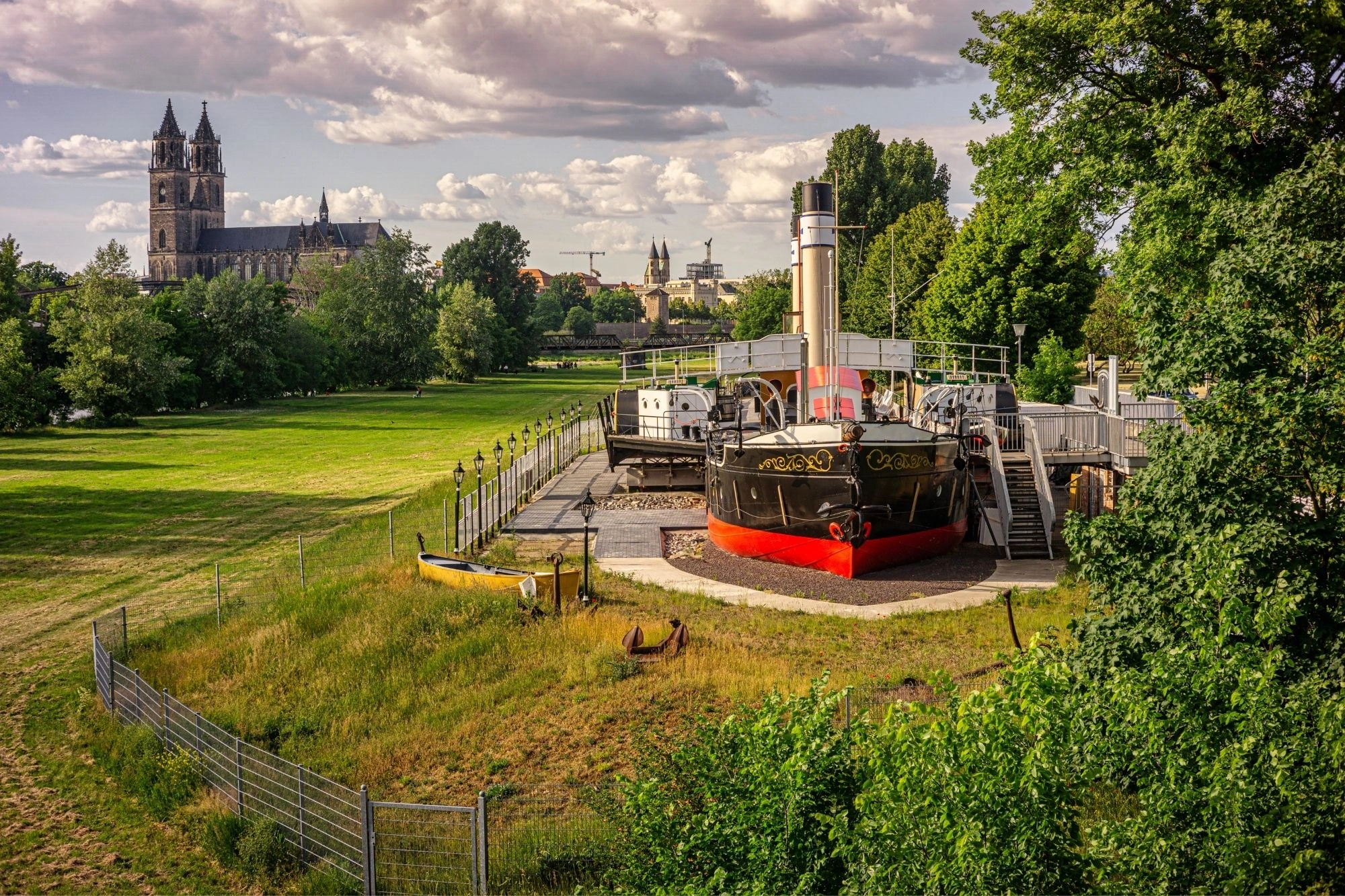 Historisches Dampfschiff auf Land in einem Park mit grünen Wiesen und Bäumen, im Hintergrund eine große Kathedrale und Stadtgebäude unter bewölktem Himmel