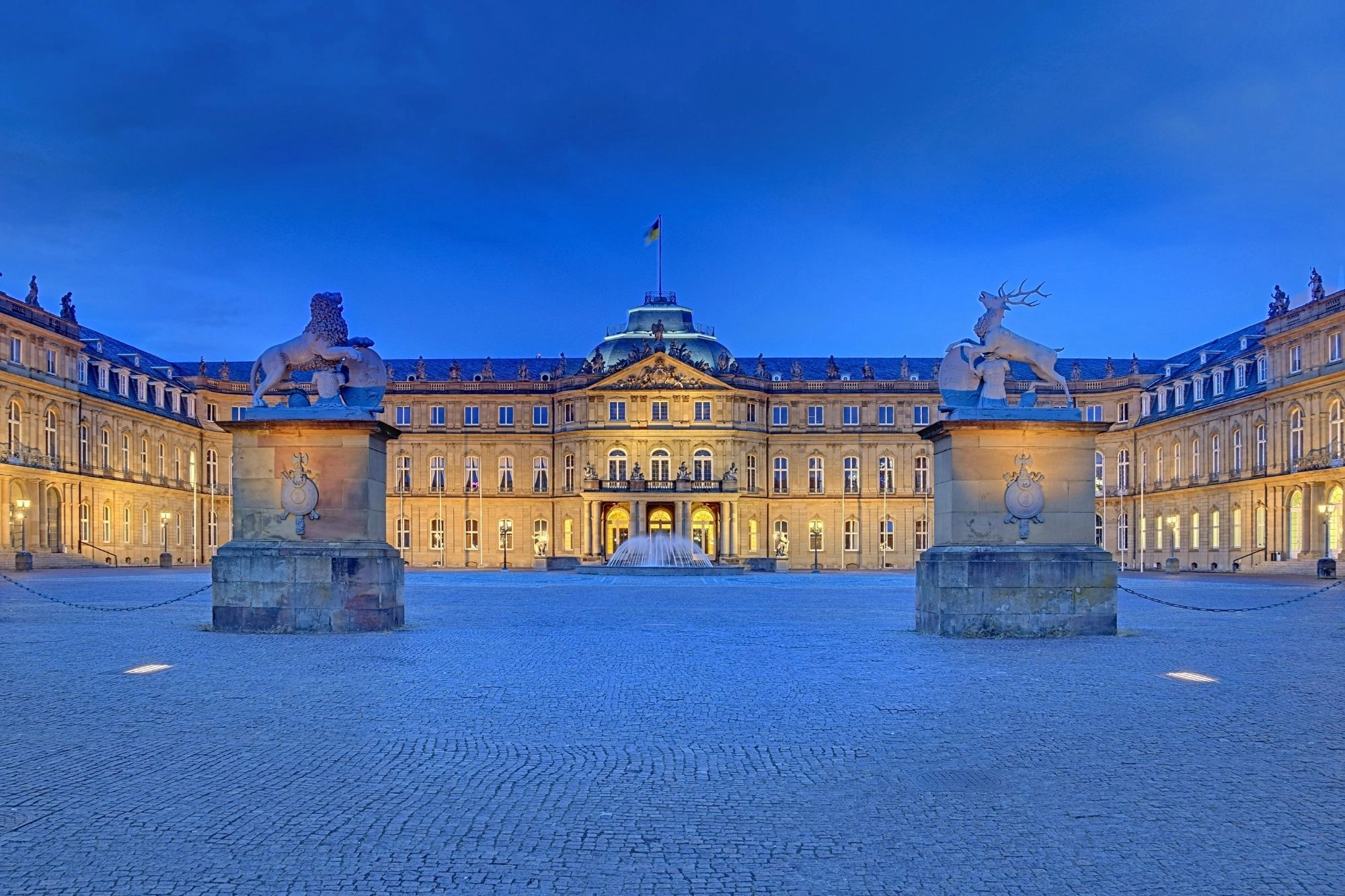 Beleuchtetes Schloss mit zwei steinernen Skulpturen eines Löwen und eines Hirsches im Vordergrund bei Abenddämmerung