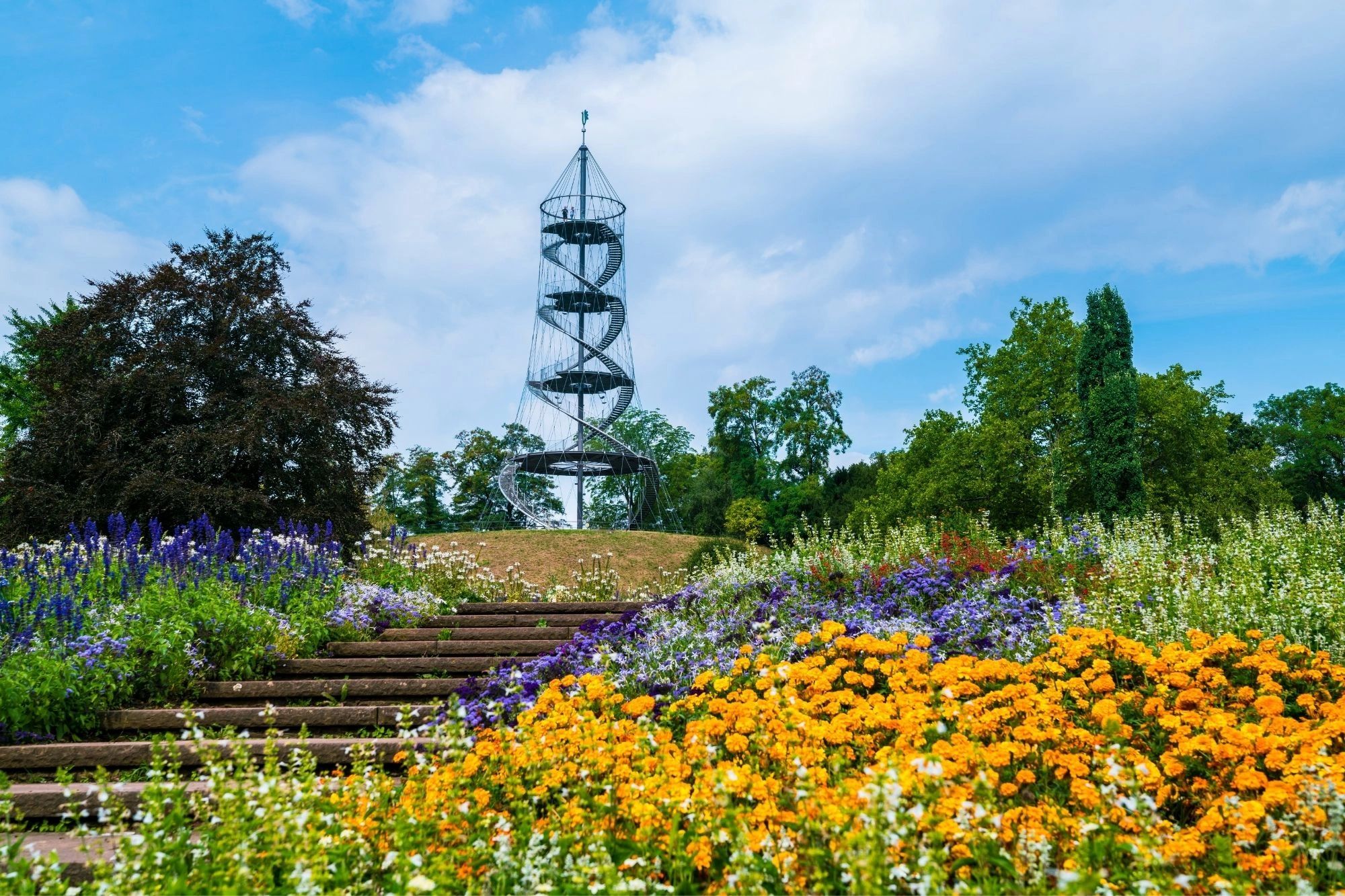 Metallspiralturm auf einem Hügel mit bunten Blumen und grünen Bäumen unter blauem Himmel