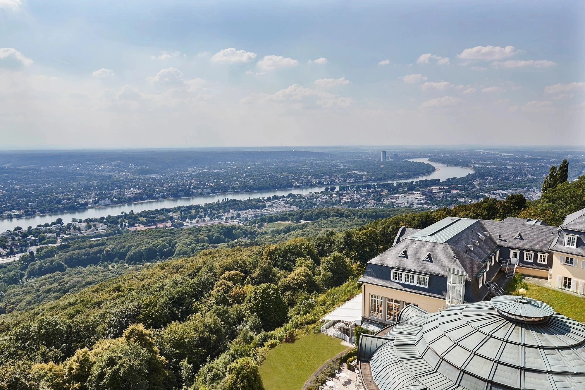 Der Ausblick vom Steigenberger Grandhotel Petersberg bietet eine beeindruckende Panoramasicht auf den Rhein, der sich majestätisch durch die Landschaft schlängelt. Die umliegenden grünen Hügel und die Stadtlandschaft erstrecken sich weit in die Ferne, während das elegante Hotelgebäude im Vordergrund den luxuriösen Charakter der Lage unterstreicht. Ein atemberaubender Blick, der Ruhe und Weite vermittelt.