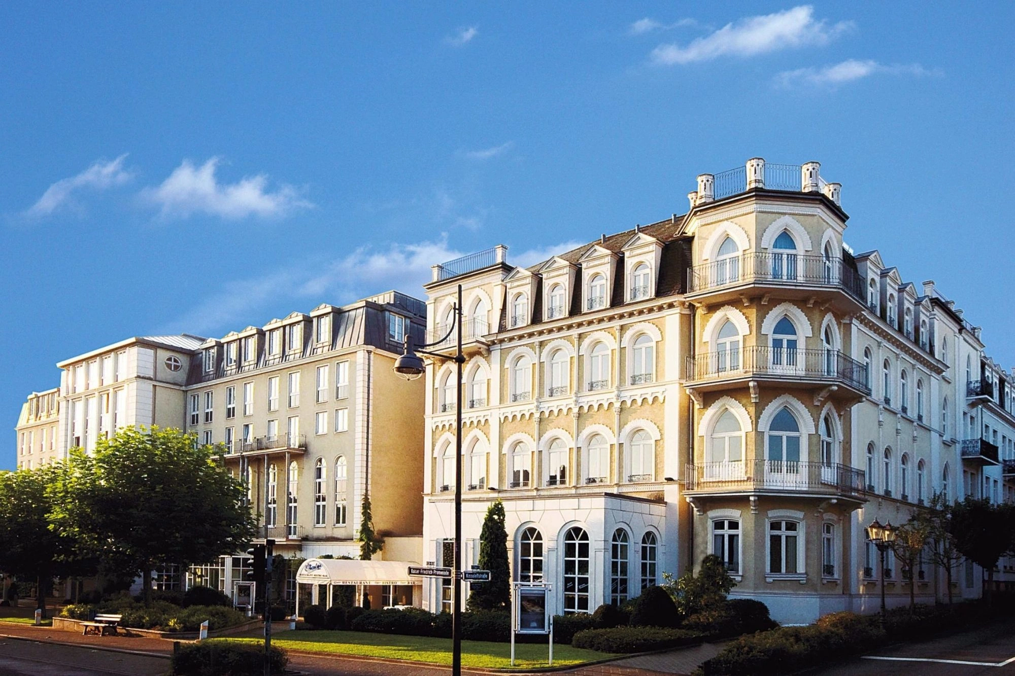 Die Außenansicht des Steigenberger Hotels in Bad Homburg zeigt eine beeindruckende, historische Fassade mit großen Fenstern und eleganten Balkonen. Das Gebäude besticht durch seine klassizistische Architektur mit Rundbögen und detailreichen Verzierungen, die dem Hotel einen majestätischen Charme verleihen. Die Fassade erstrahlt im Sonnenlicht, und die gepflegte Umgebung mit Bäumen unterstreicht die ruhige, luxuriöse Atmosphäre des Hotels.