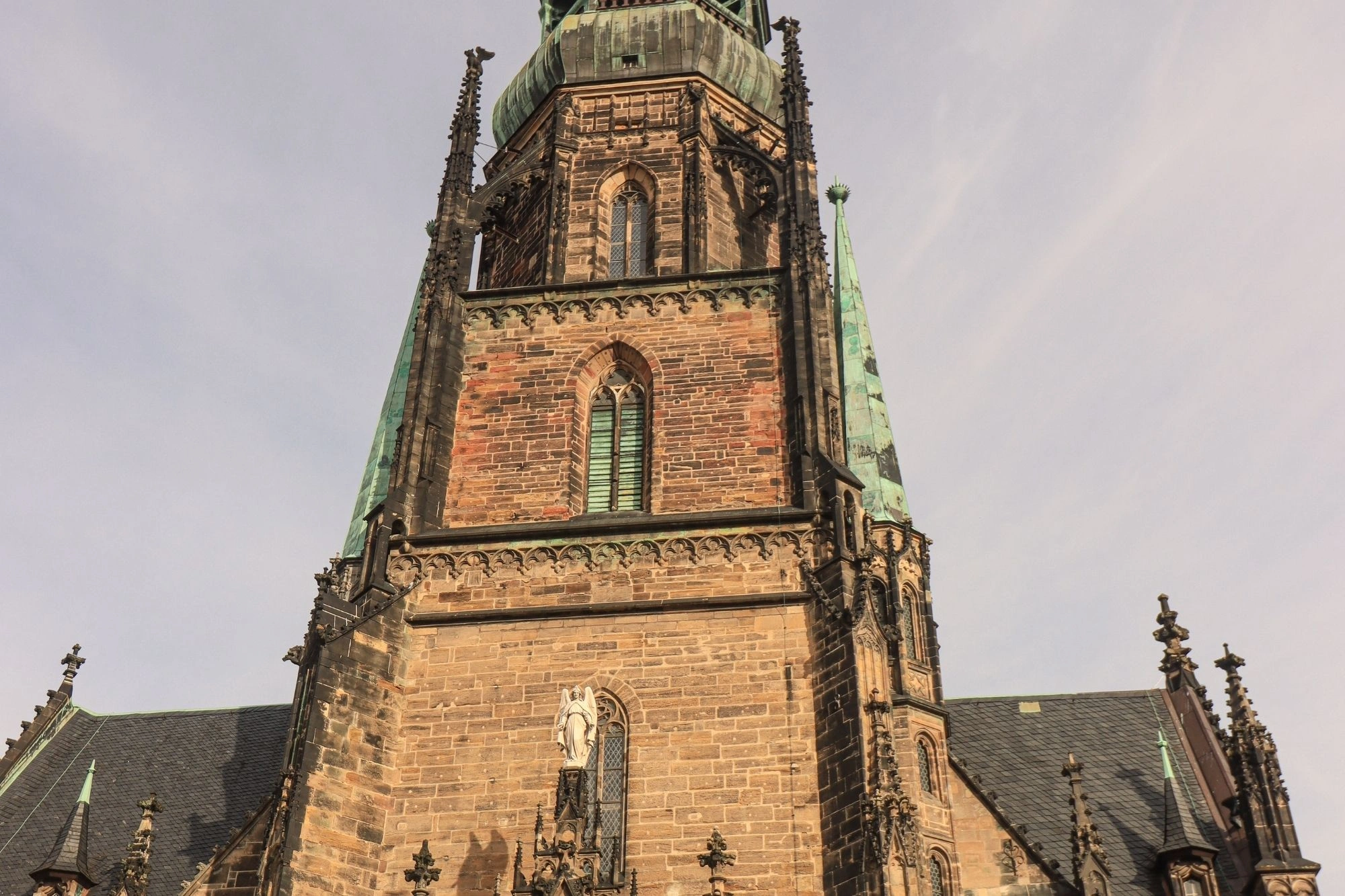 Gotischer Kirchturm mit spitzem Dach und mehreren Fenstern aus braunem Stein unter bewölktem Himmel.