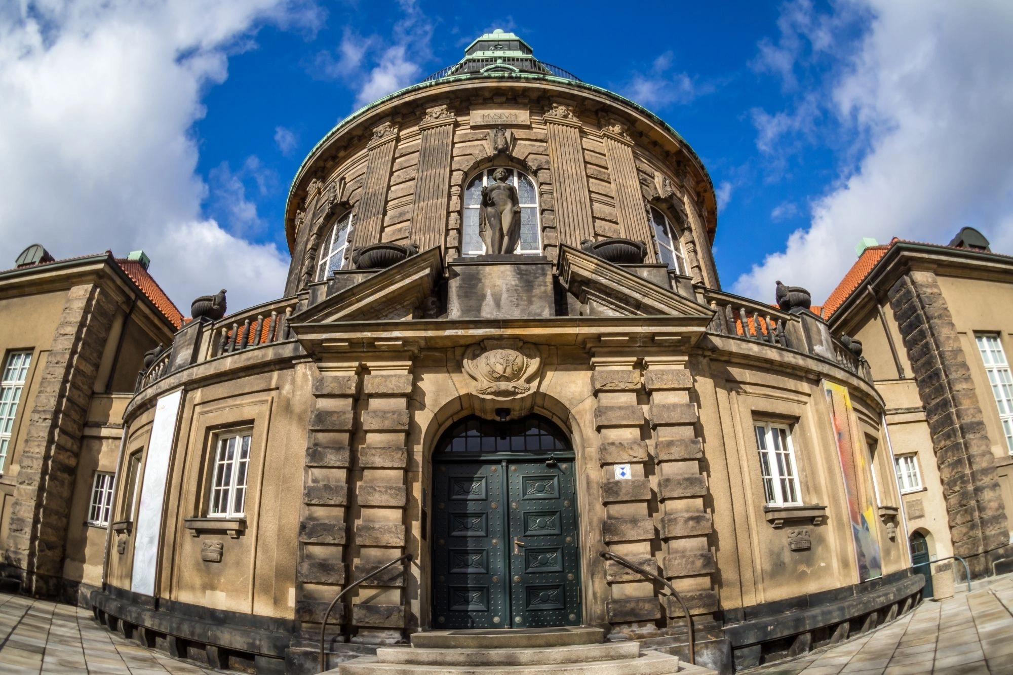 Rundes historisches Gebäude mit steinernen Verzierungen, einer Statue in einer Nische über dem Eingang und einem großen dunklen Holztor.