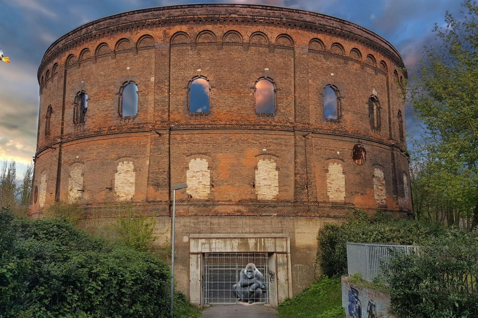 Rundes, altes Backsteingebäude mit hohen Fenstern, umgeben von Büschen und Bäumen.