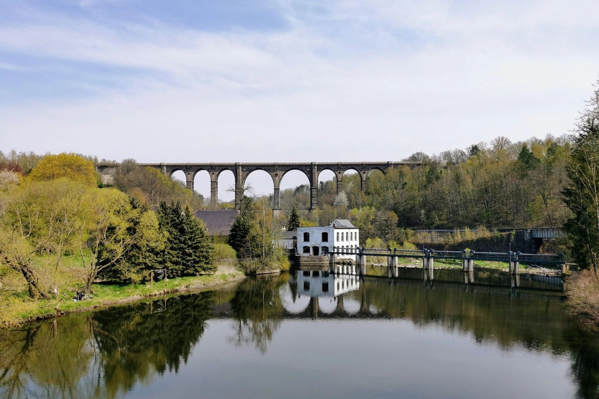 Ein weißes Gebäude am Ufer eines ruhigen Sees, im Hintergrund ein hoher Eisenbahnviadukt mit mehreren Bögen, umgeben von Bäumen und leicht bewölktem Himmel.