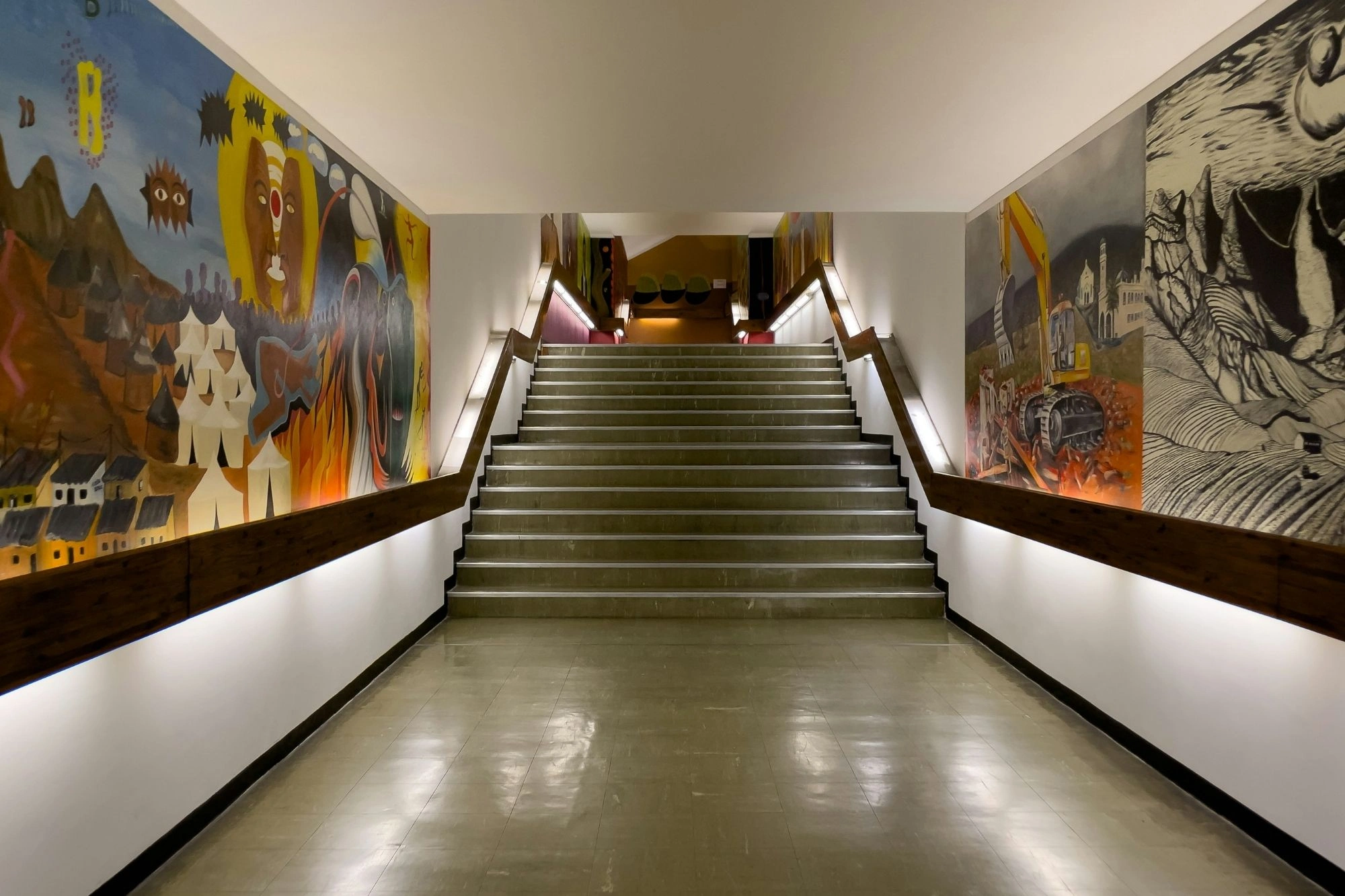 Treppenaufgang mit bunten Wandgemälden auf beiden Seiten, darunter abstrakte und figürliche Motive, beleuchtet von Wandlampen.