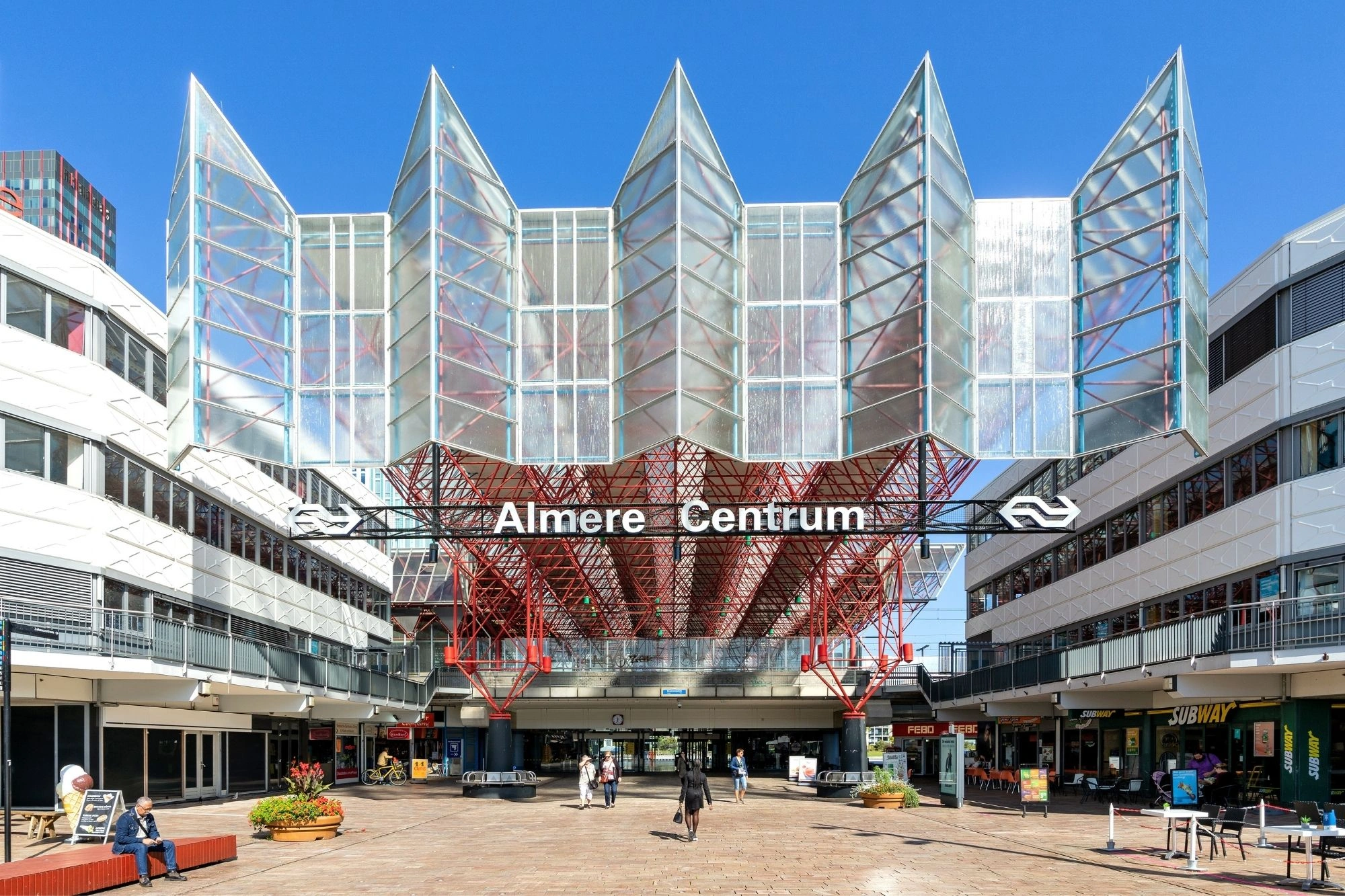 Eingang des Bahnhofs Almere Centrum mit auffälliger Glas- und Stahlkonstruktion, rotem Dach und umliegenden Geschäften bei klarem Himmel.