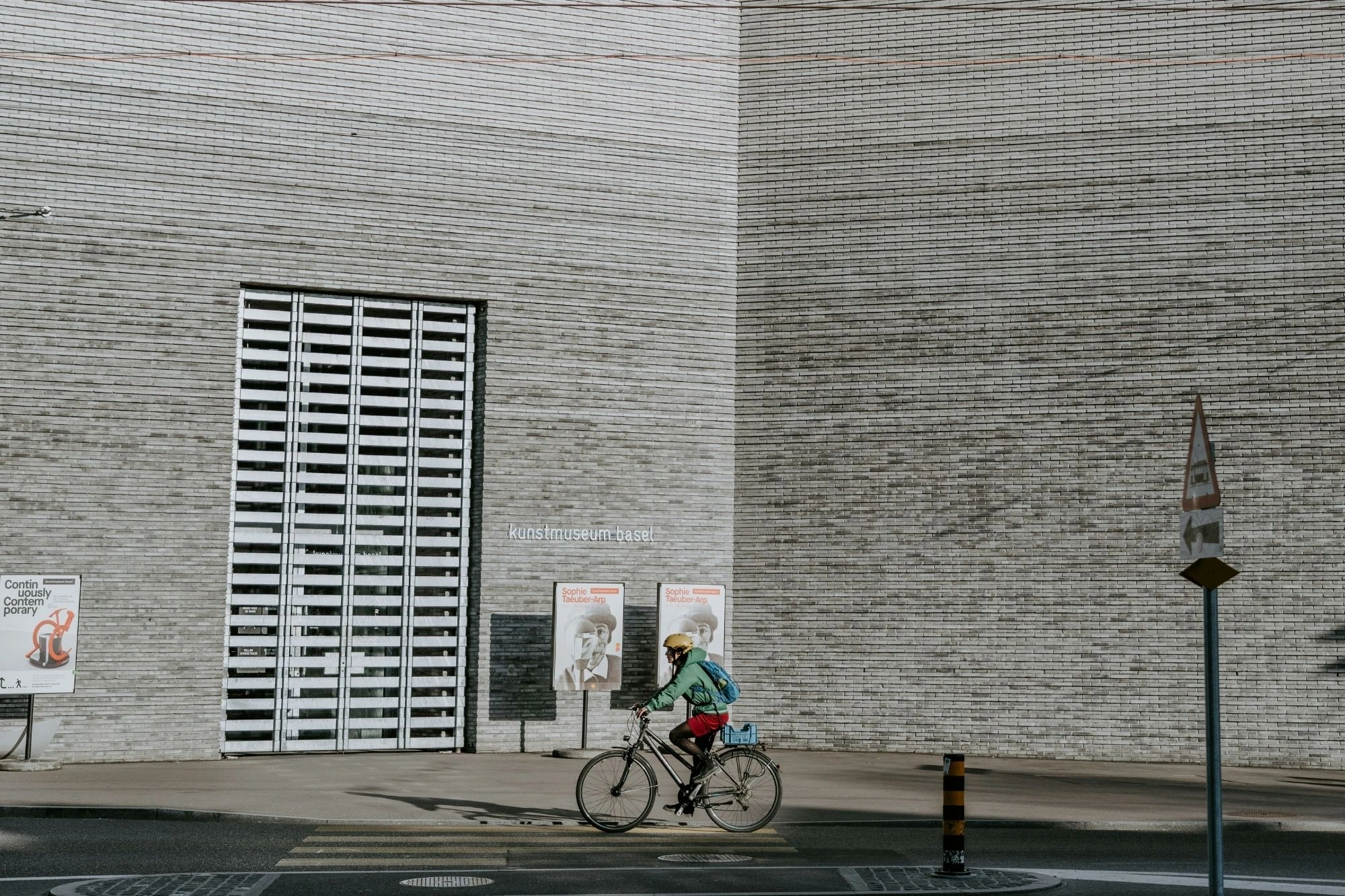 Fahrradfahrerin mit Helm, roter Hose und blauem Rucksack vor einer großen, hellen Backsteinmauer mit zwei Plakaten und einem großen Gitterfenster.