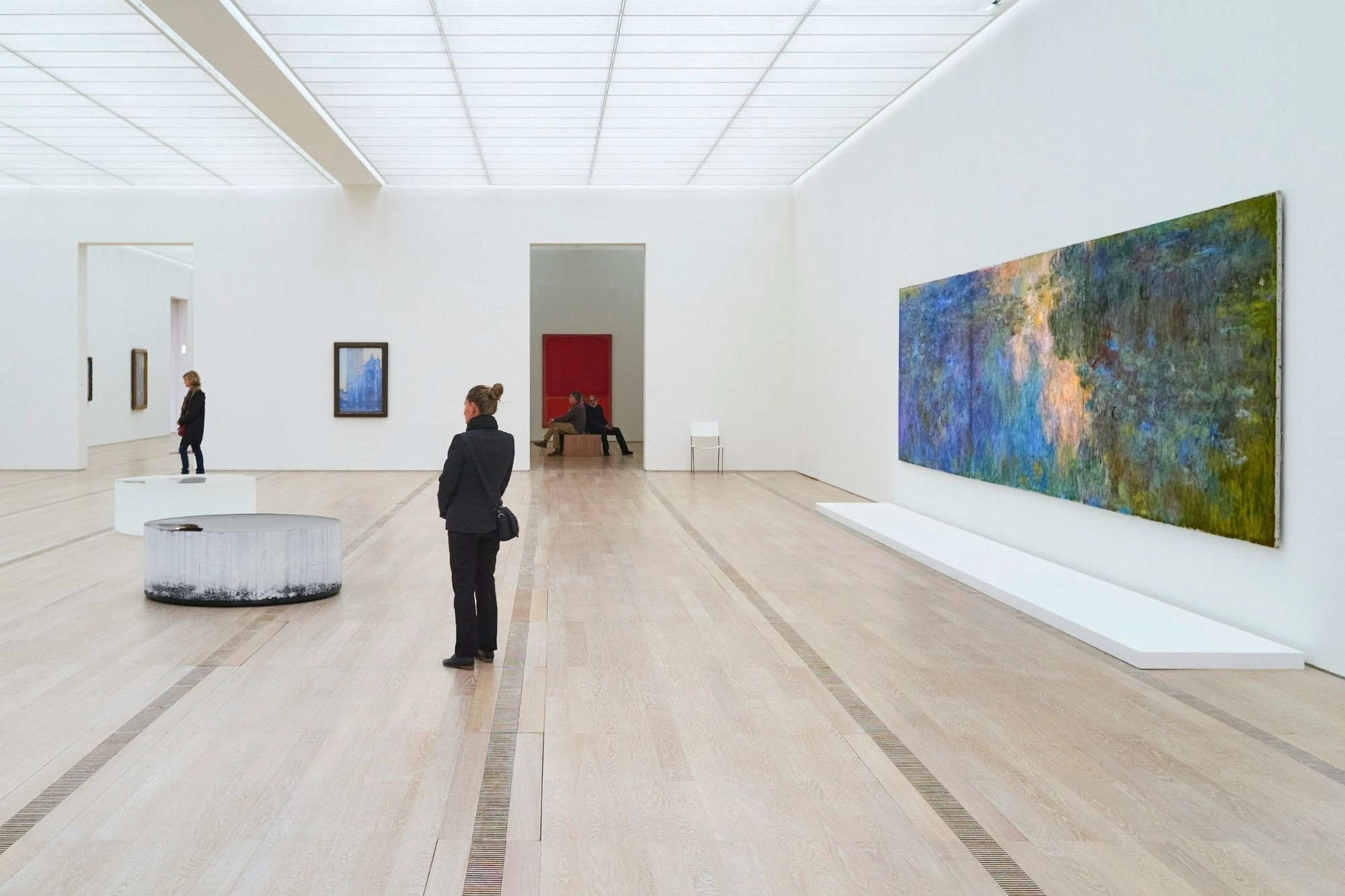 Großes abstraktes Gemälde mit Blau- und Grüntönen an weißer Wand in modernem Museum, zwei Personen betrachten Kunstwerke, eine sitzt auf Bank im Hintergrund.