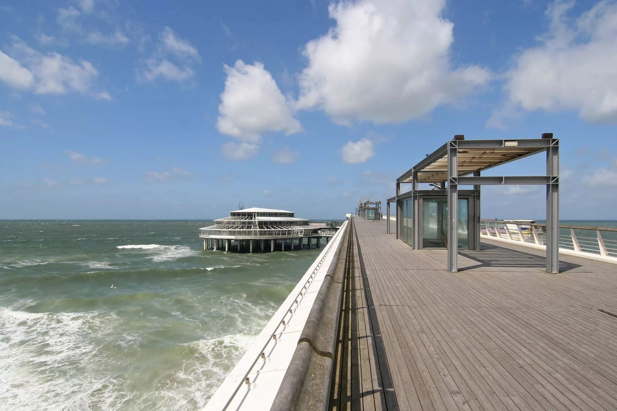 Pier Restaurant Scheveningen Ein Panorama des Pier Restaurants in Scheveningen, aufgenommen bei hellem Tageslicht. Die Wellen schlagen sanft gegen die Säulen des Piers und erzeugen eine beruhigende Geräuschkulisse. Die Sonne reflektiert sich auf dem Wasser und betont die einladende Atmosphäre des Lokals. Dieses Bild vermittelt eine entspannte, maritime Stimmung und lädt zum Verweilen ein.