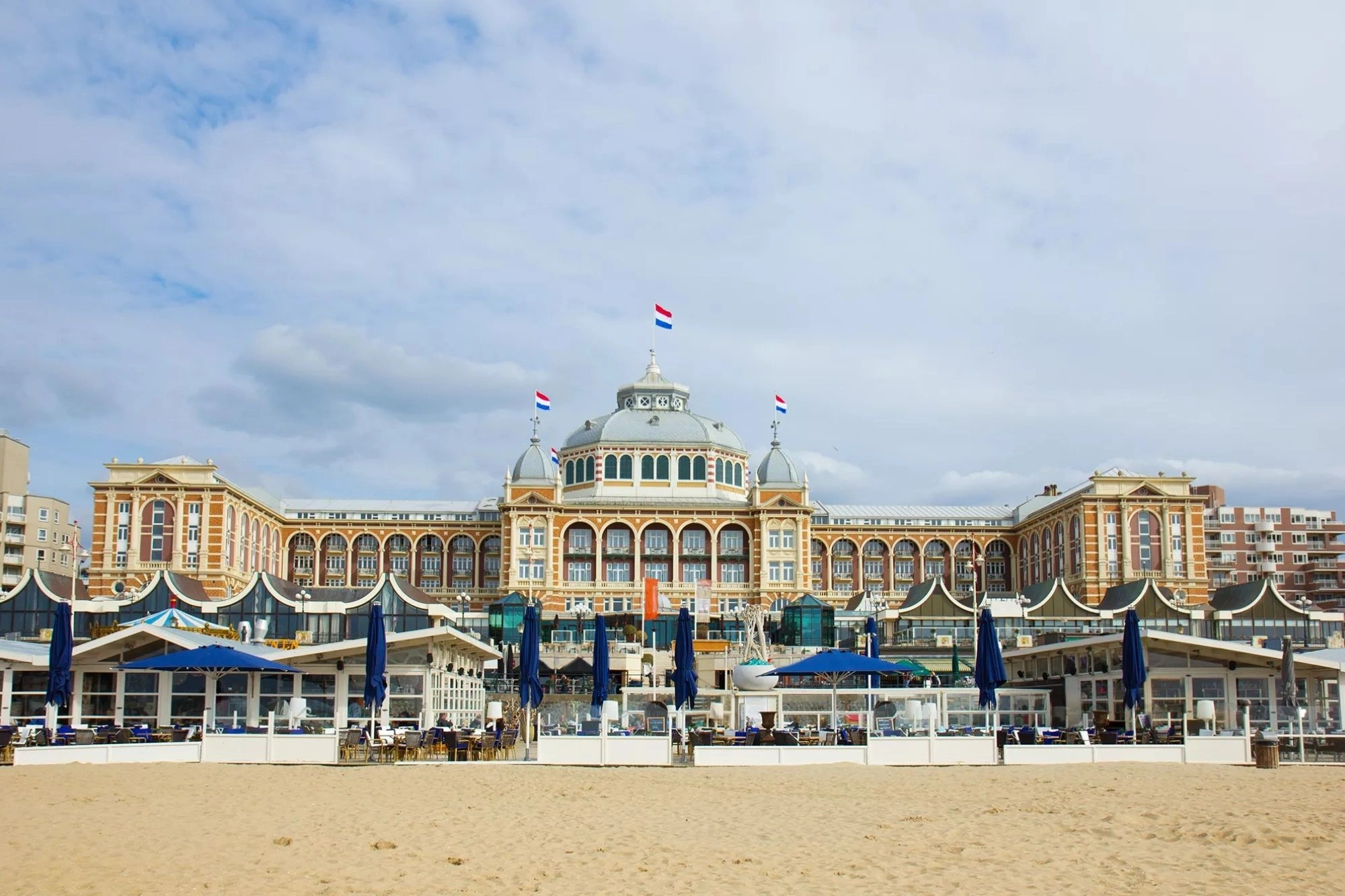 Das Kurhaus in Scheveningen mit seiner einladenden Außenterrasse, auf der einige Menschen verweilen. Bei Tageslicht strahlt das Gebäude eine majestätische Atmosphäre aus, während der nahegelegene Strand eine entspannte Kulisse bietet. Die harmonische Verbindung von Architektur und Küstenumgebung lädt zu einem genussvollen Aufenthalt ein.