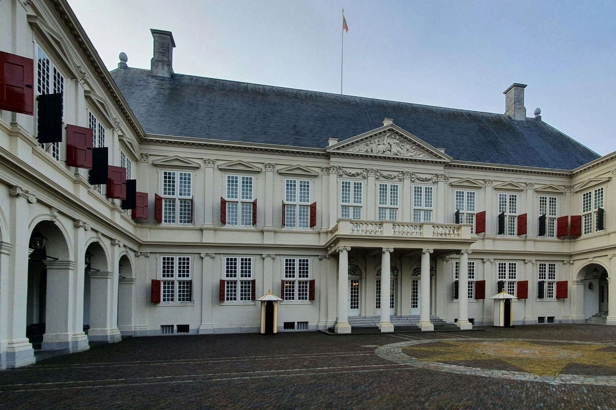 Fassade eines historischen Gebäudes mit weißen Wänden, roten Fensterläden und einem steinernen Dachgiebel unter bewölktem Himmel.