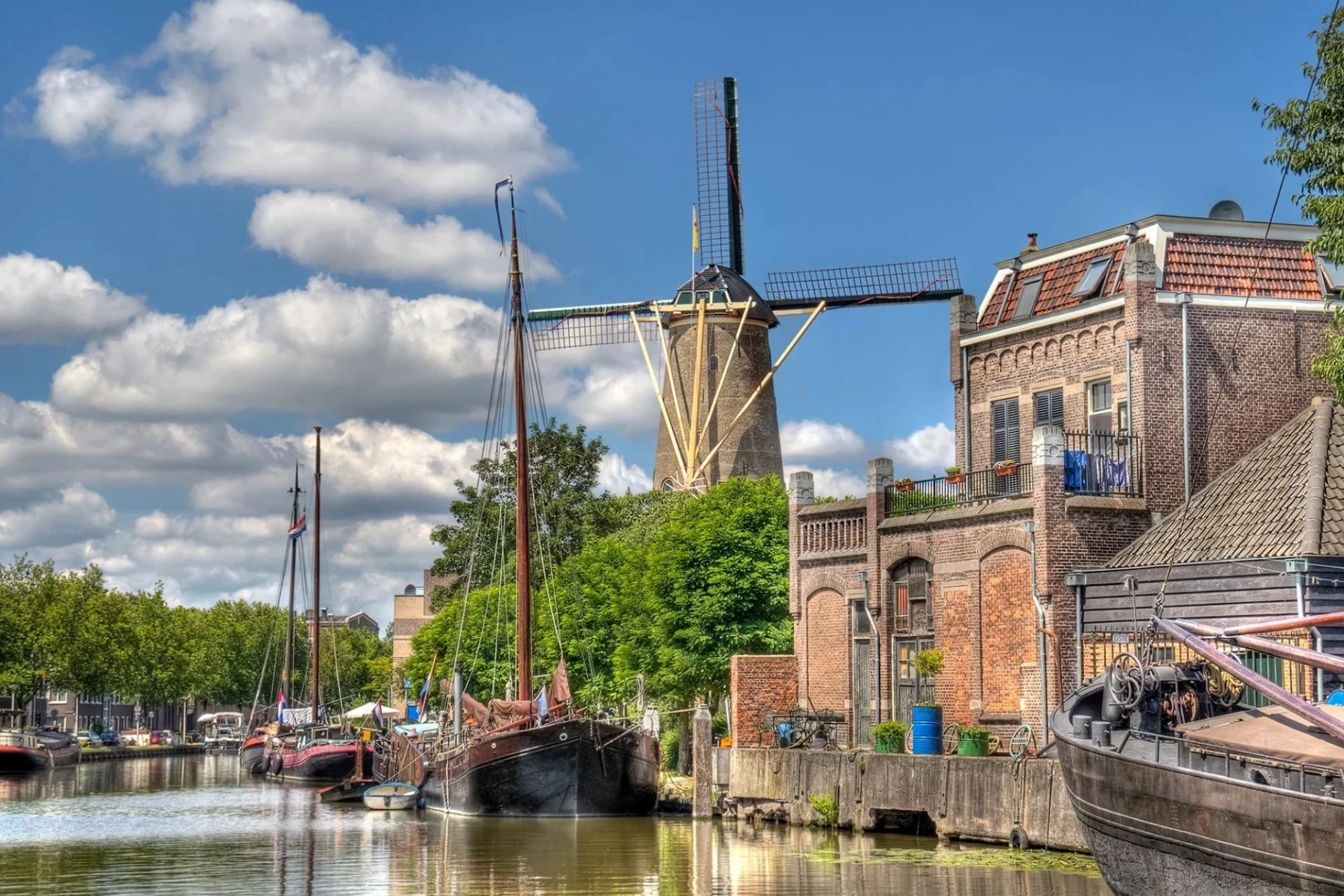 Eine malerische Windmühle vor einer Gracht, umgeben von angelegten Booten und Schiffen, bei klarem Wetter. Die reflektierende Wasseroberfläche verstärkt die Schönheit der Szene, während die lebhaften Farben und der strahlend blaue Himmel eine einladende Atmosphäre schaffen. Dieses Bild fängt die harmonische Verbindung von traditioneller Architektur und maritimem Leben ein.