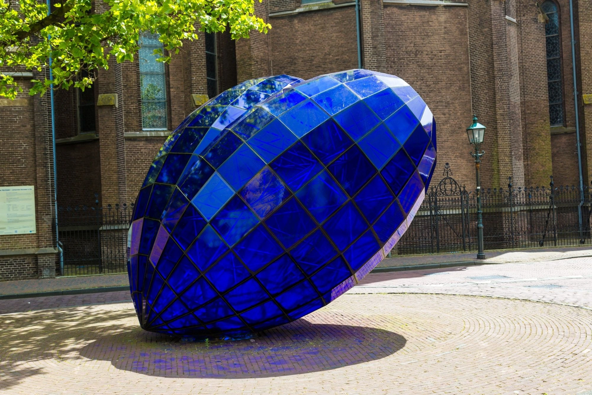 Große blaue Skulptur in Form eines facettierten Herzens aus Glas oder Keramik vor Backsteinmauer und Laterne.