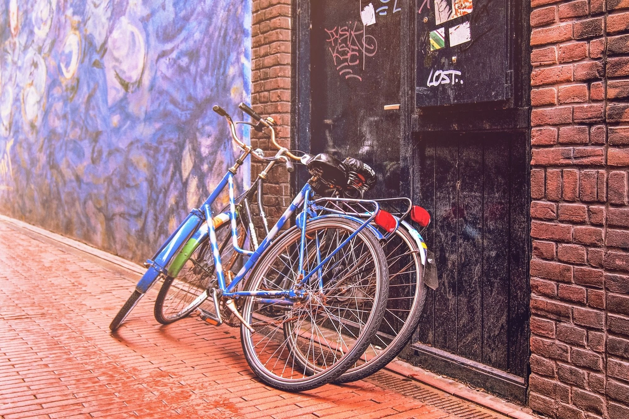 Zwei Fahrräder lehnen an einer Backsteinmauer neben einer schwarzen Tür mit Graffiti.