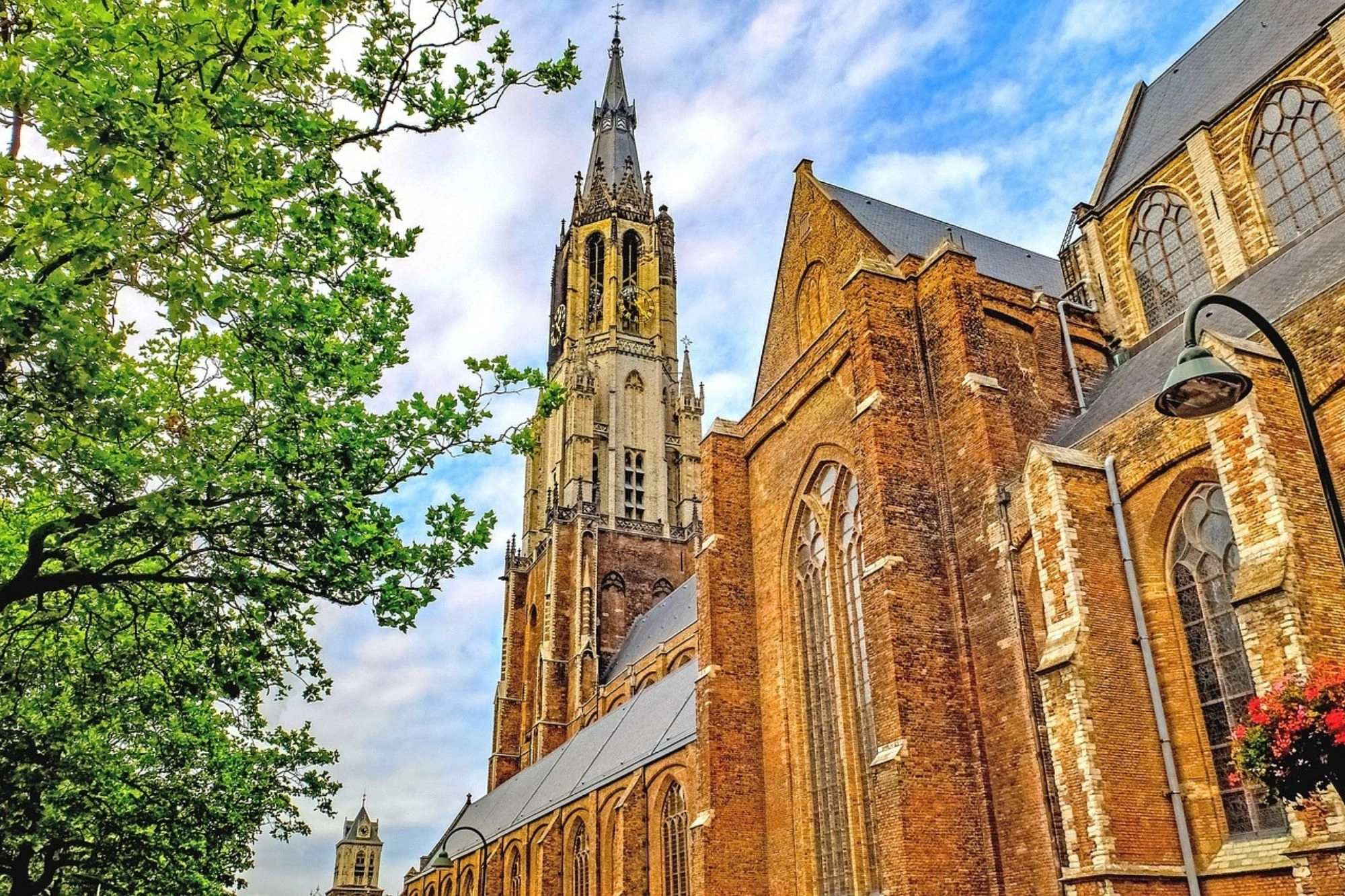 Gotische Backsteinkirche mit hohem Turm und spitzem Dach, umgeben von grünem Laub und blauem Himmel mit Wolken.