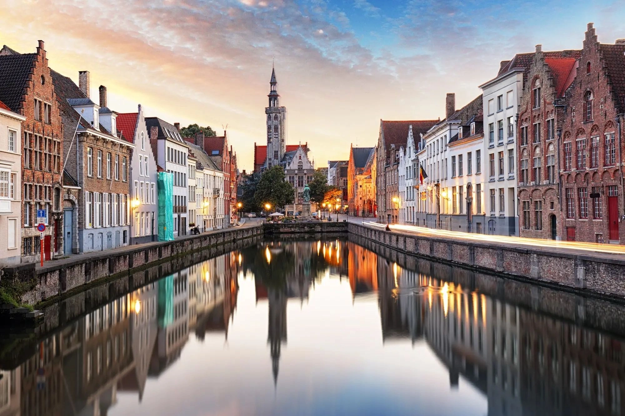 Blick von der Brücke auf den Kanal entlang der Jan van Eyckplein-Statue bei Abenddämmerung, mit Stadtbeleuchtung und Häuserzeilen, die sich im Wasser spiegeln.
