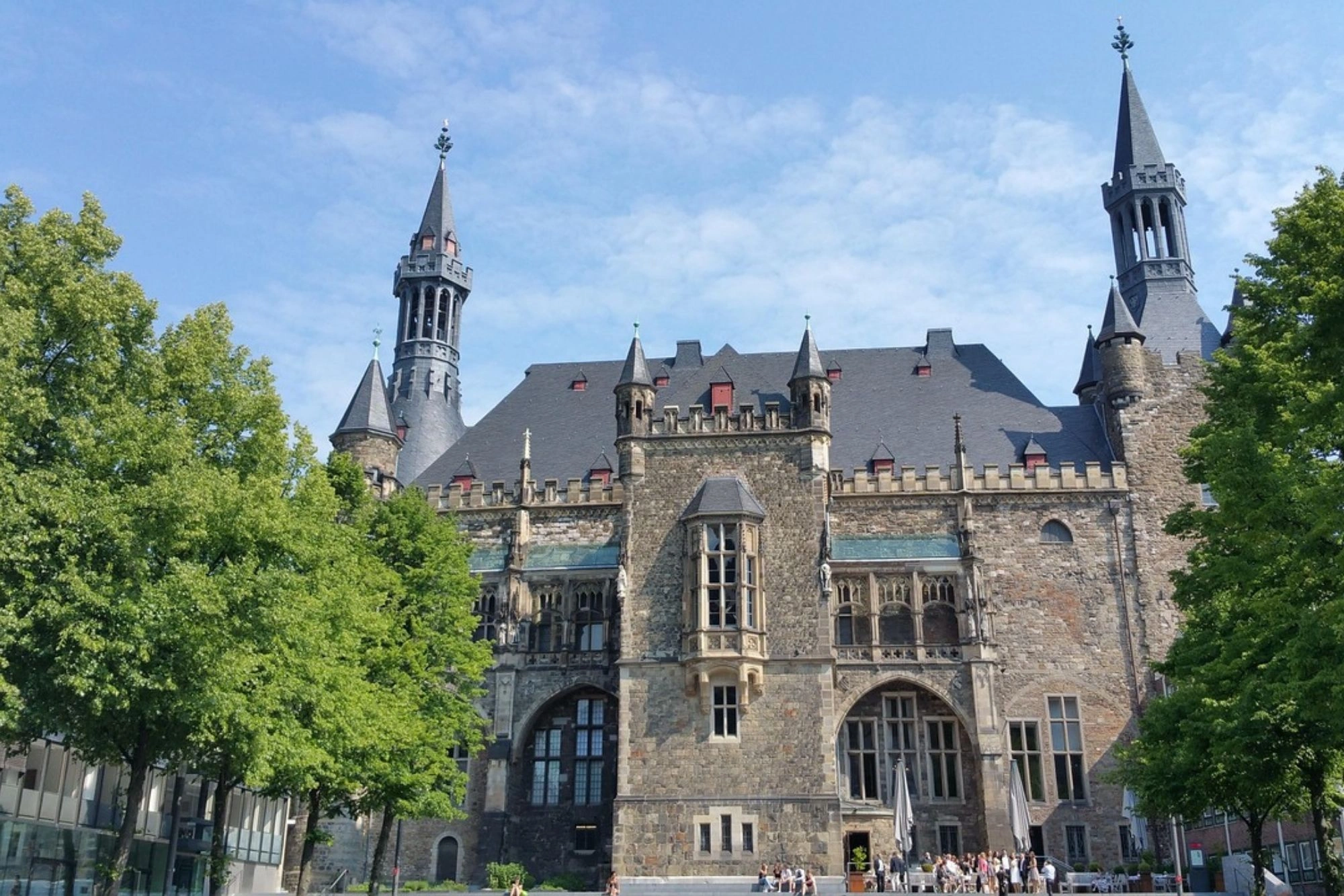 Historisches Schloss mit mehreren Türmen und steinernen Fassaden, umgeben von grünen Bäumen unter blauem Himmel.