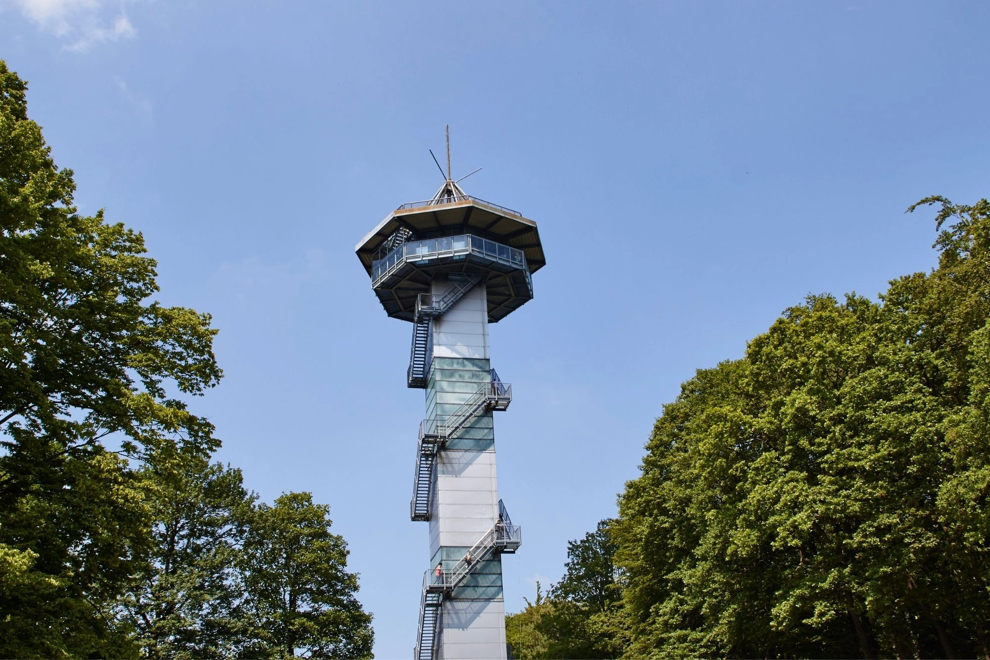 Aussichtsturm mit umlaufender Außentreppe, umgeben von grünen Bäumen unter blauem Himmel.